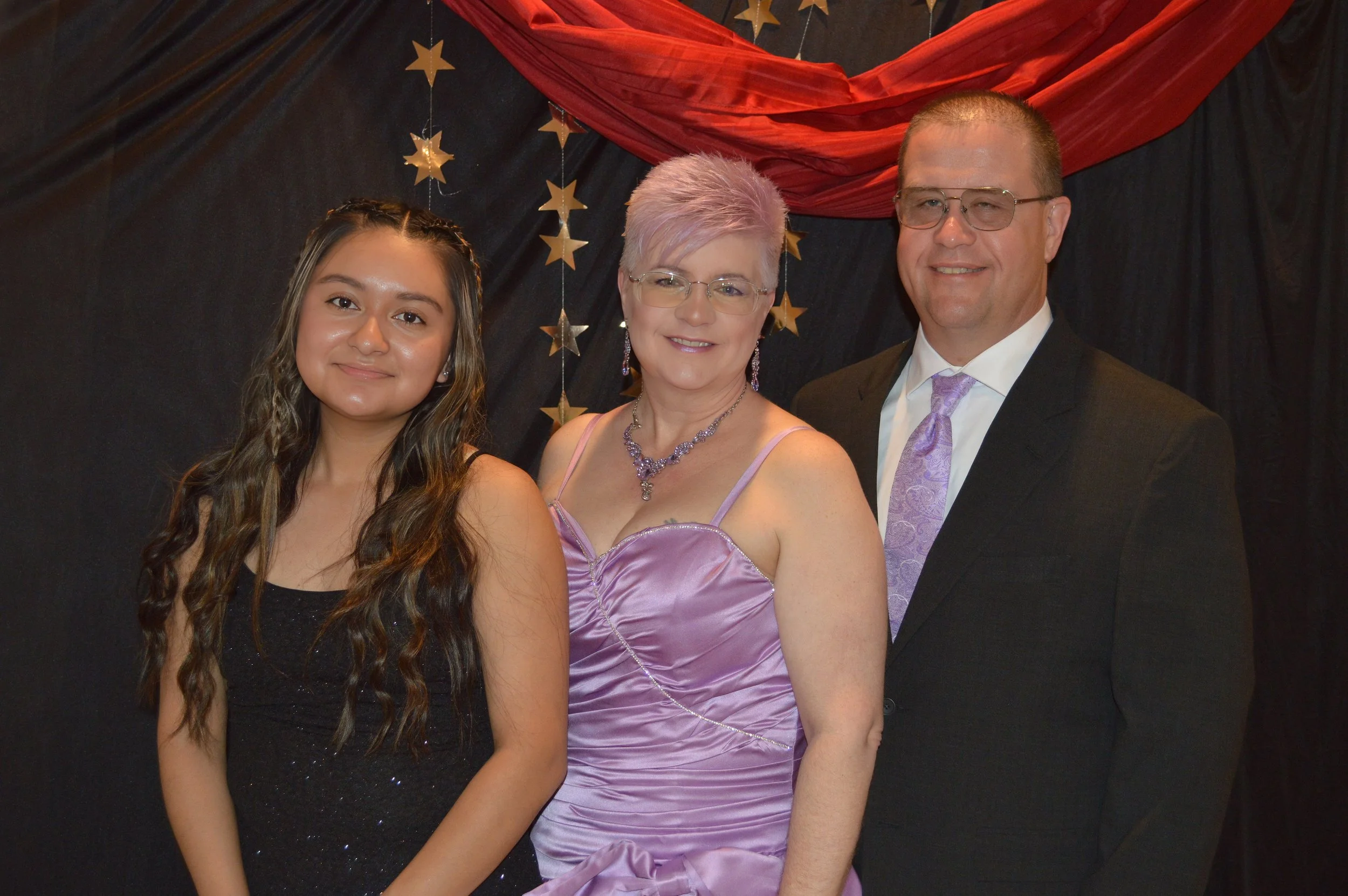 A young woman, a middle-aged woman, and a man are dressed in formal attire, standing close together and smiling. The background features black and red drapery with hanging gold star decorations.