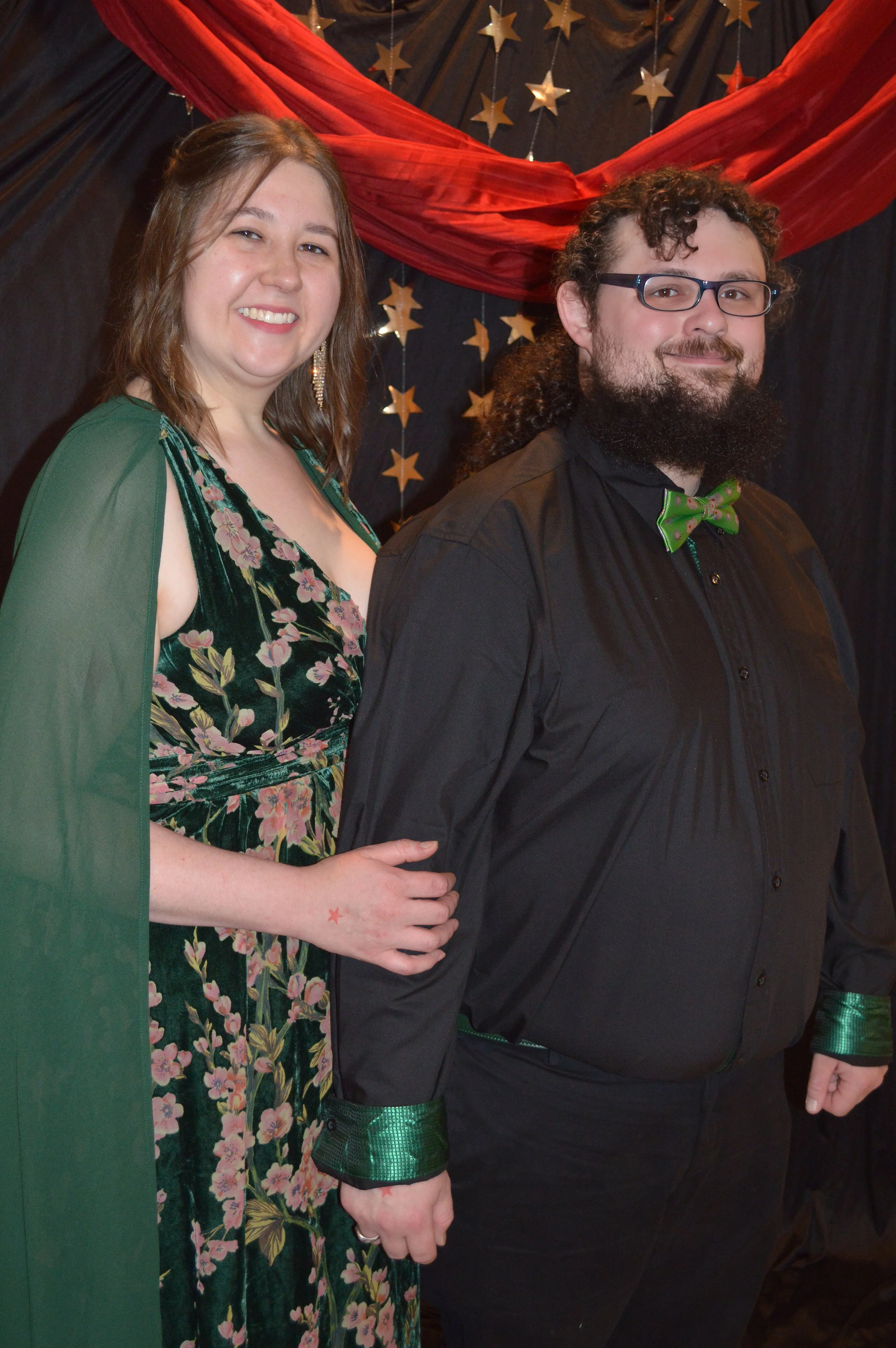 A woman and a man dressed in formal attire, standing together at a celebration or event. The woman is wearing a floral dress with a green cape, and the man is wearing a black shirt with green cuffs and a green polka dot bow tie. The background featur