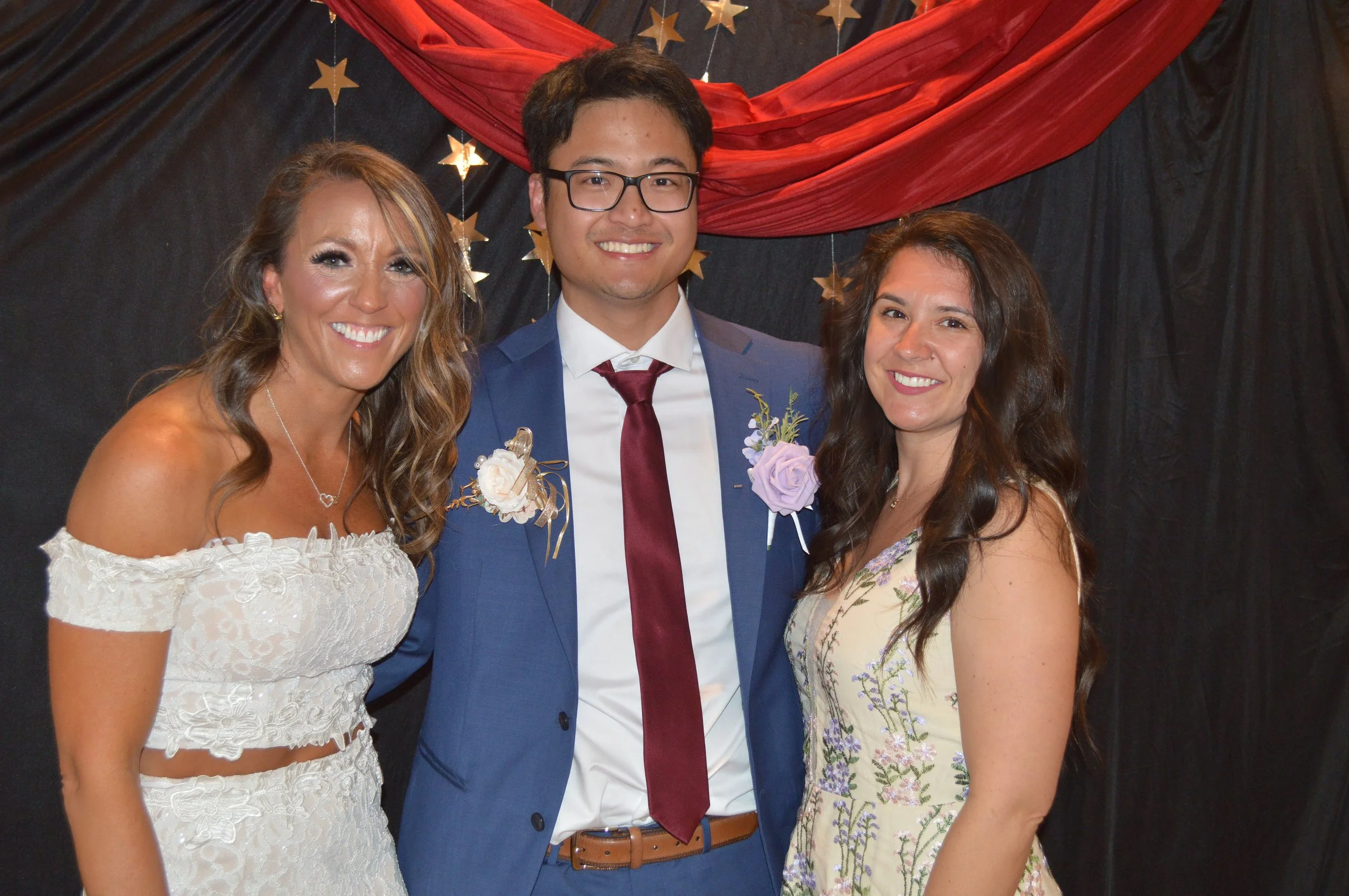 Three women and one man standing together at a celebration, with black and red drapes and gold star decorations in the background, all smiling at the camera.