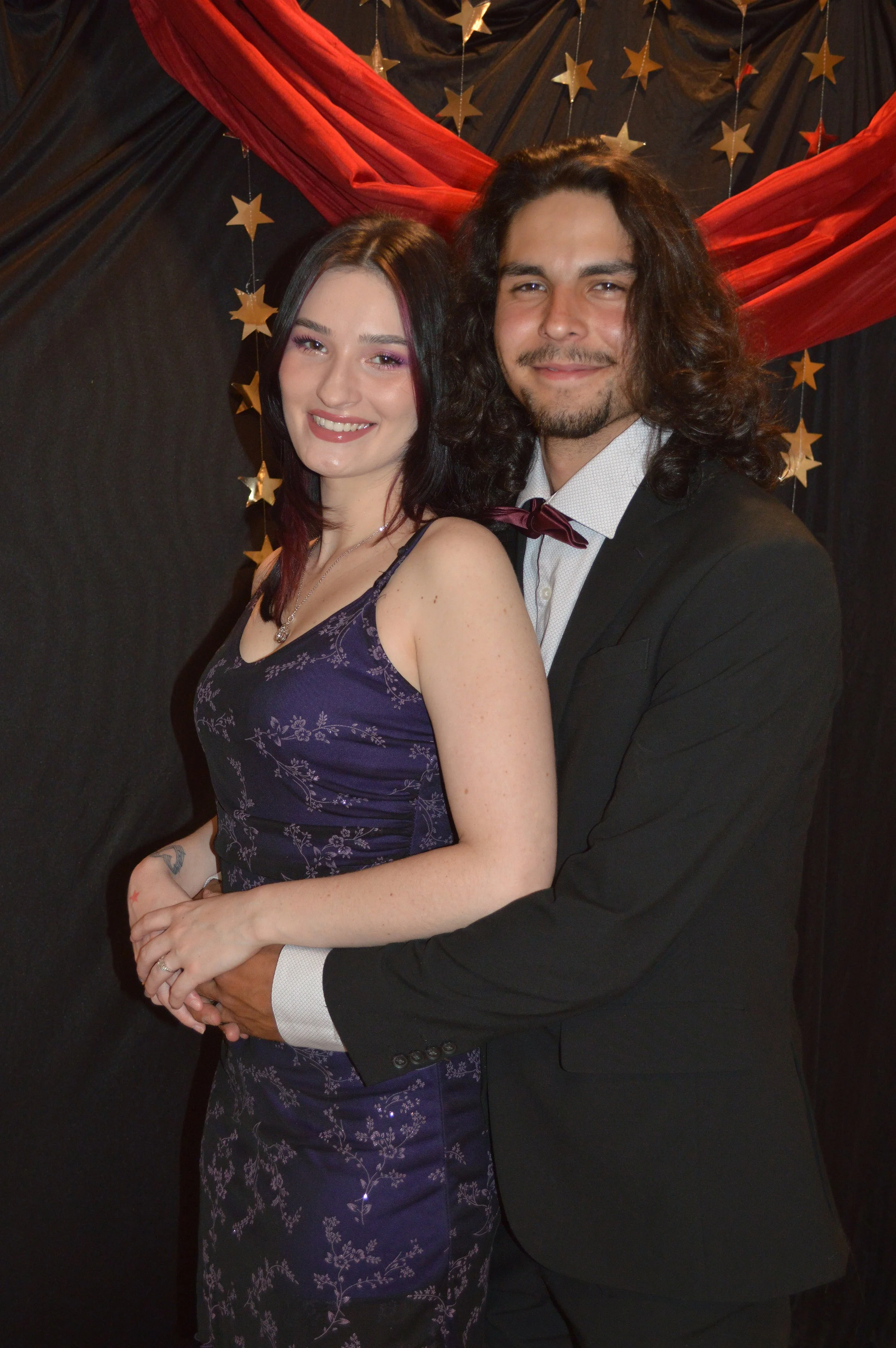 A smiling couple dressed elegantly, the woman in a purple floral dress and the man in a black suit with a white shirt and maroon bow tie, standing in front of a black backdrop decorated with red fabric and gold stars.