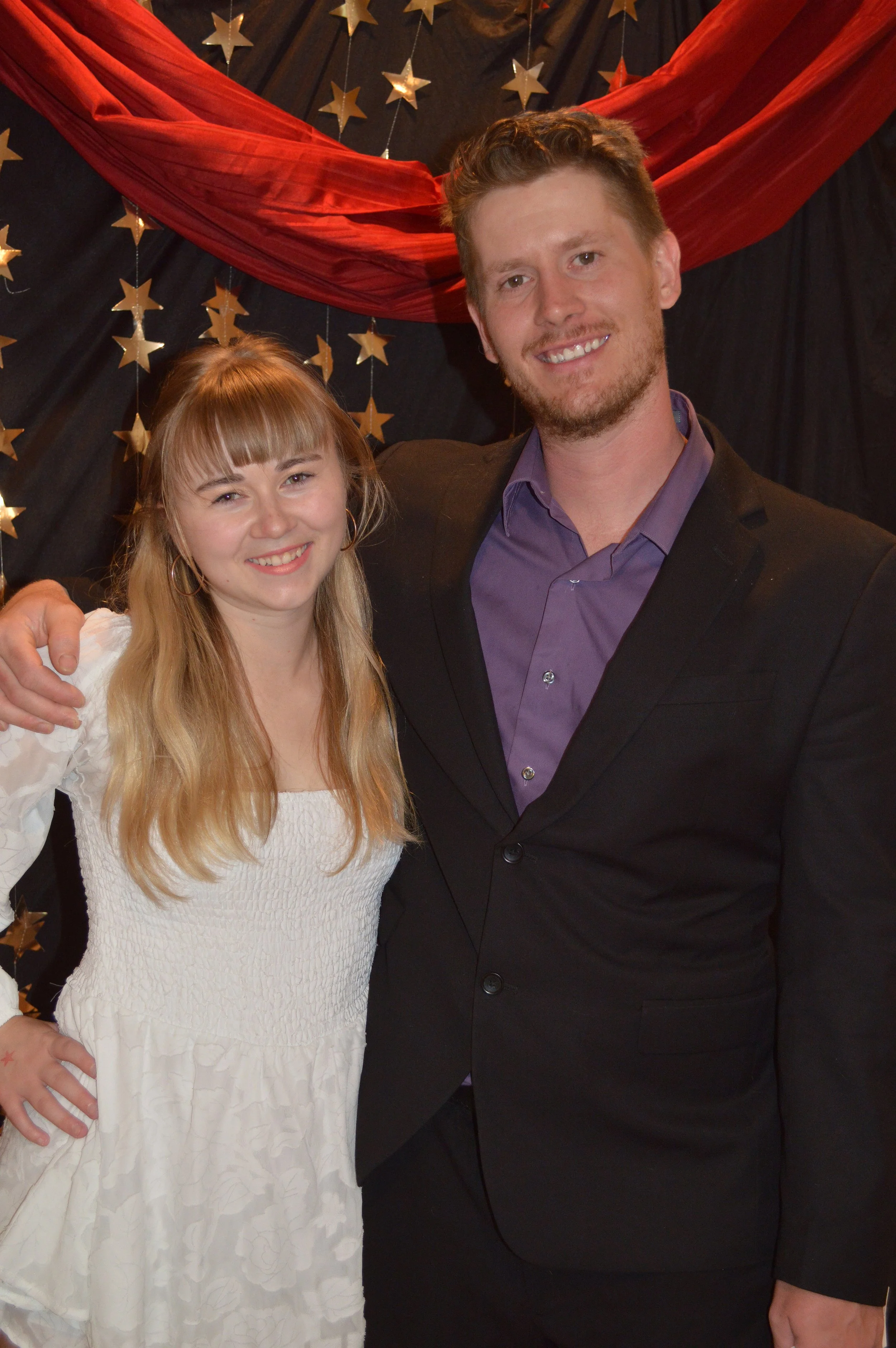 A young woman and a young man smiling and posing together at a decorated event with gold star decorations and red drapery in the background.