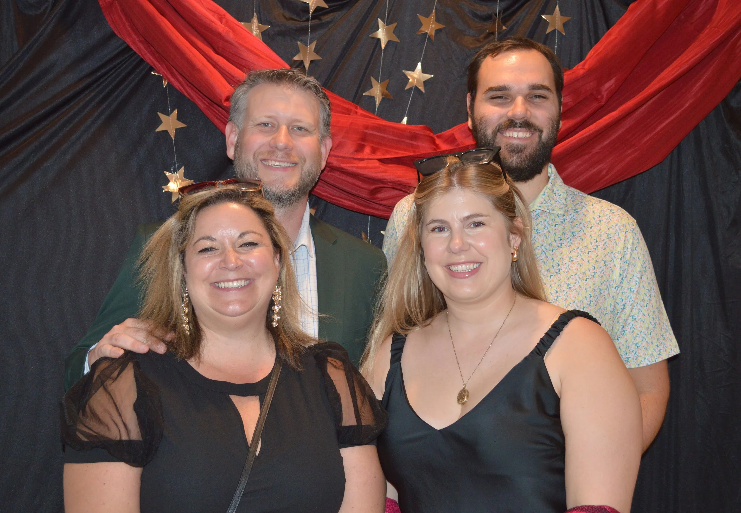 Five smiling people at a party, standing in front of a black backdrop with red fabric and gold stars, celebrating together.