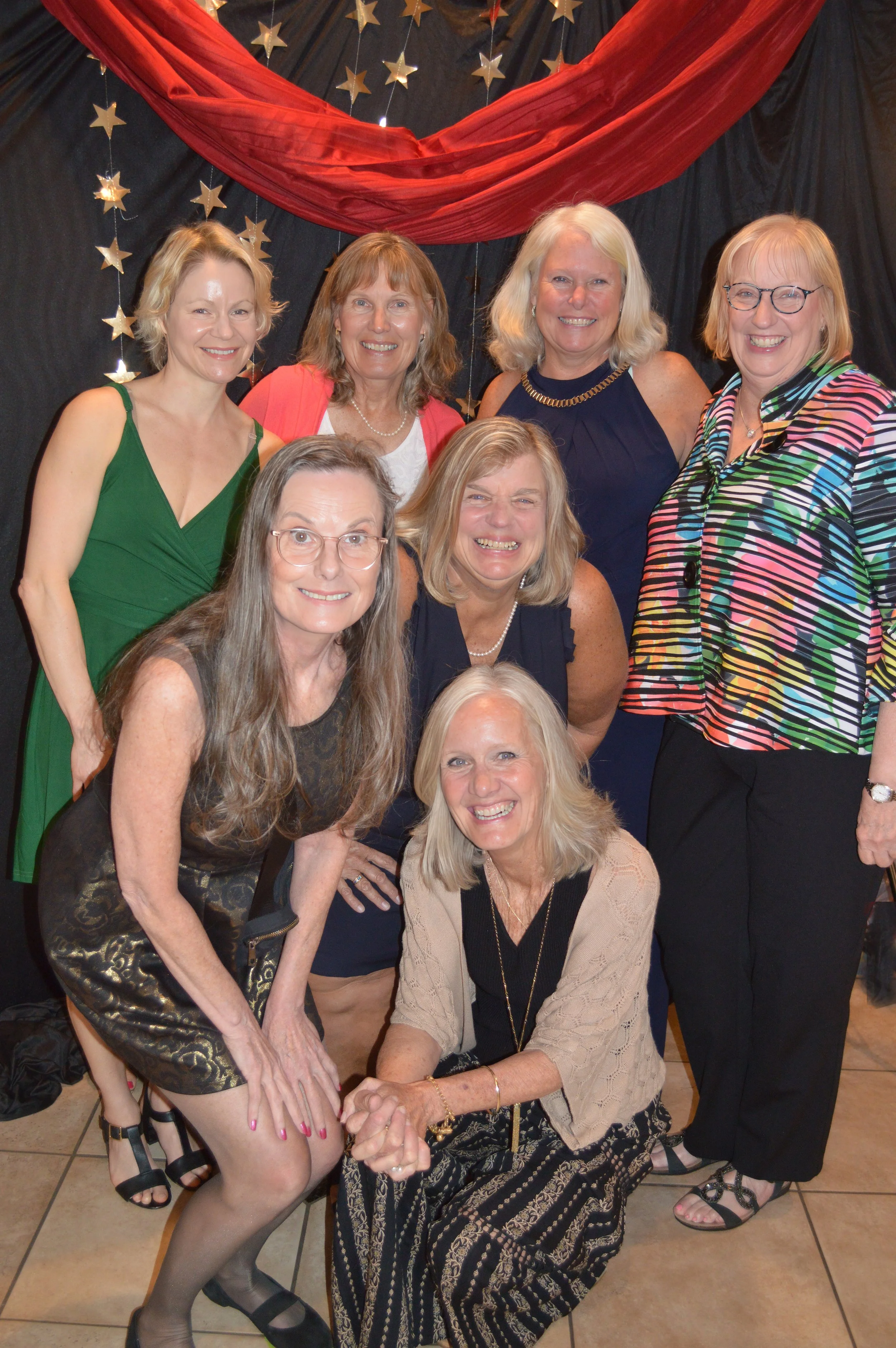 Group of nine women smiling and posing together at a celebration. The background features black and red drapes with star decorations.