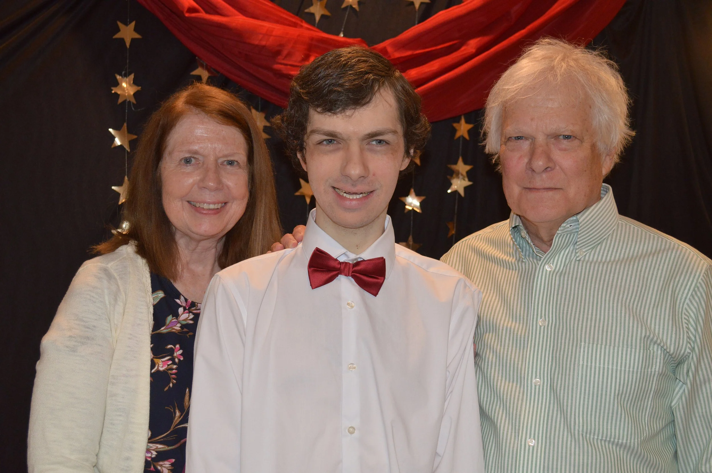 A young man in a white shirt and red bow tie standing between an older woman with red hair and an older man with white hair, all smiling, with a backdrop of black and red drapes and star decorations.