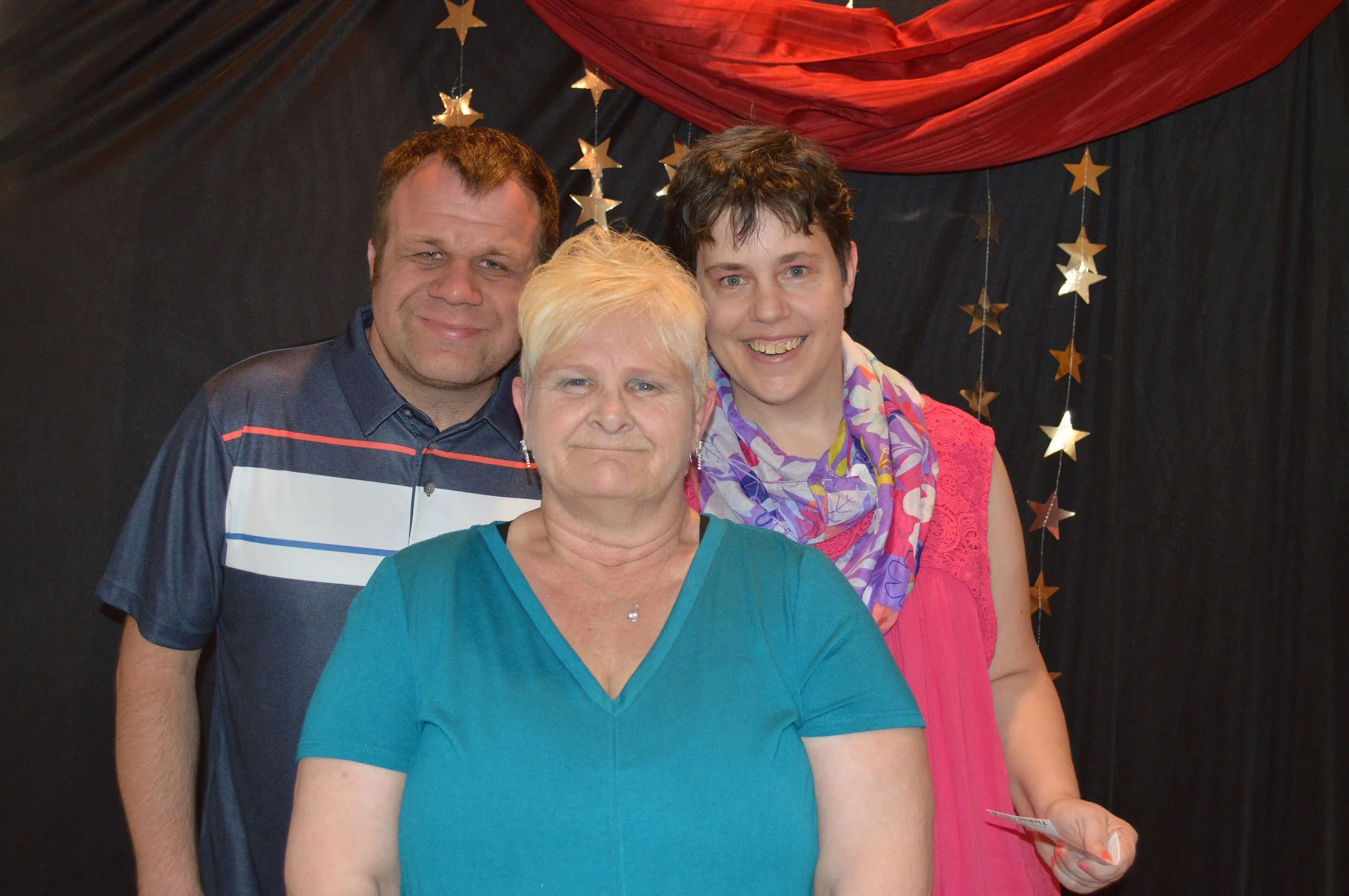 Three people, two women and one man, posing together indoors in front of a dark background with gold star decorations and red drapes.