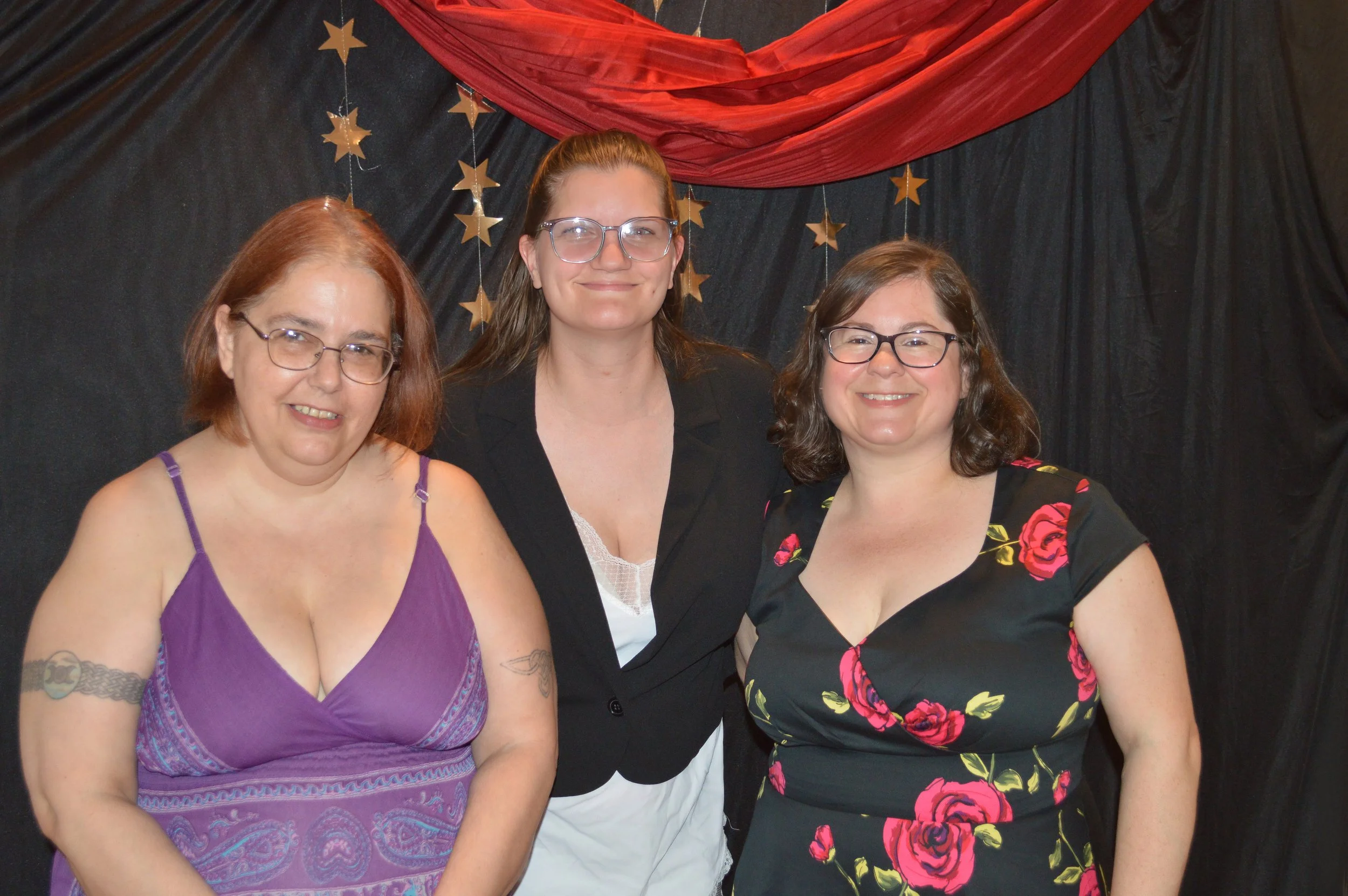 Three women standing side by side, smiling, in front of a black background decorated with gold stars and a red drape.