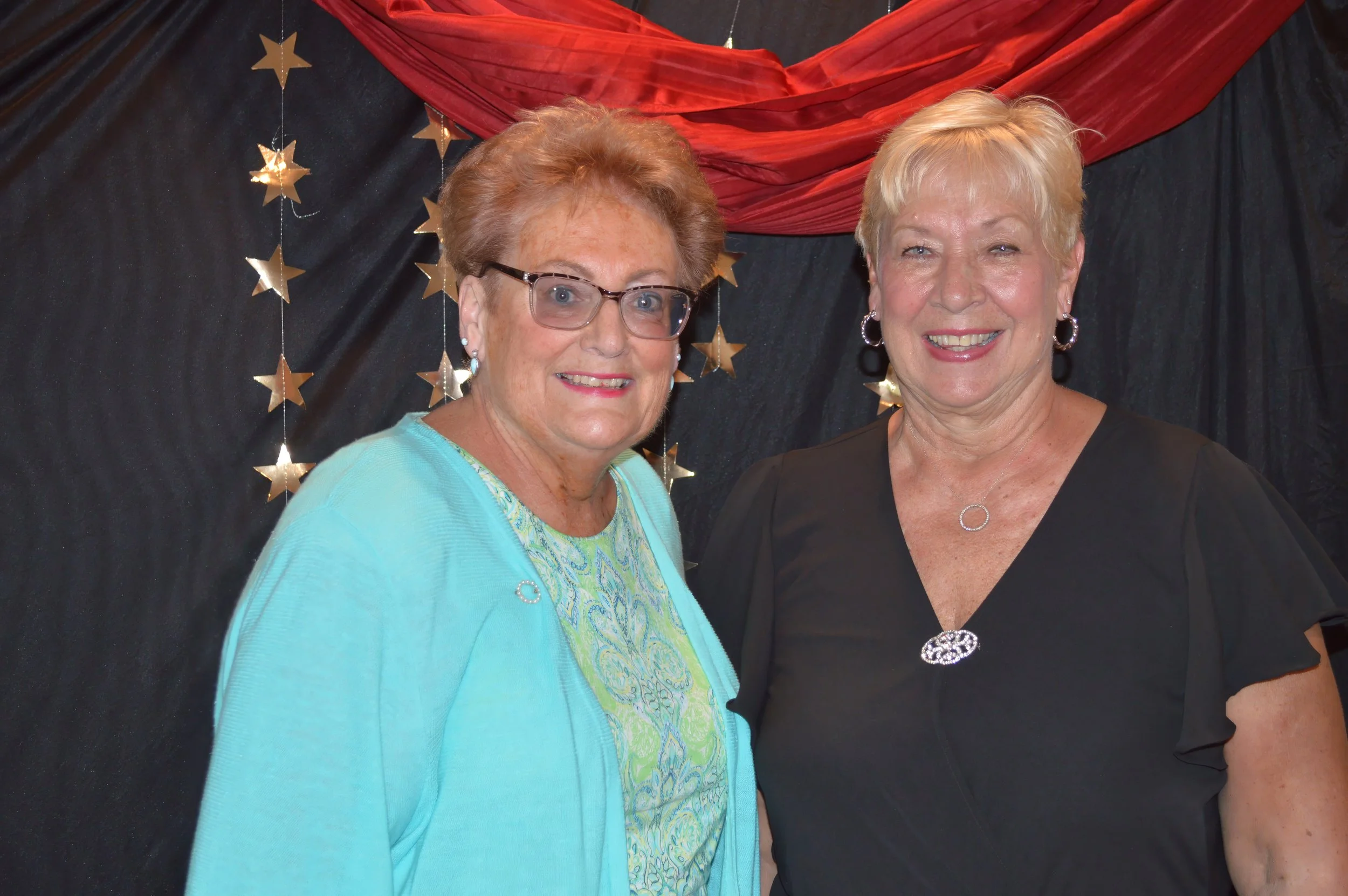 Two smiling older women standing together in front of black and red decorated backdrop with star ornaments.