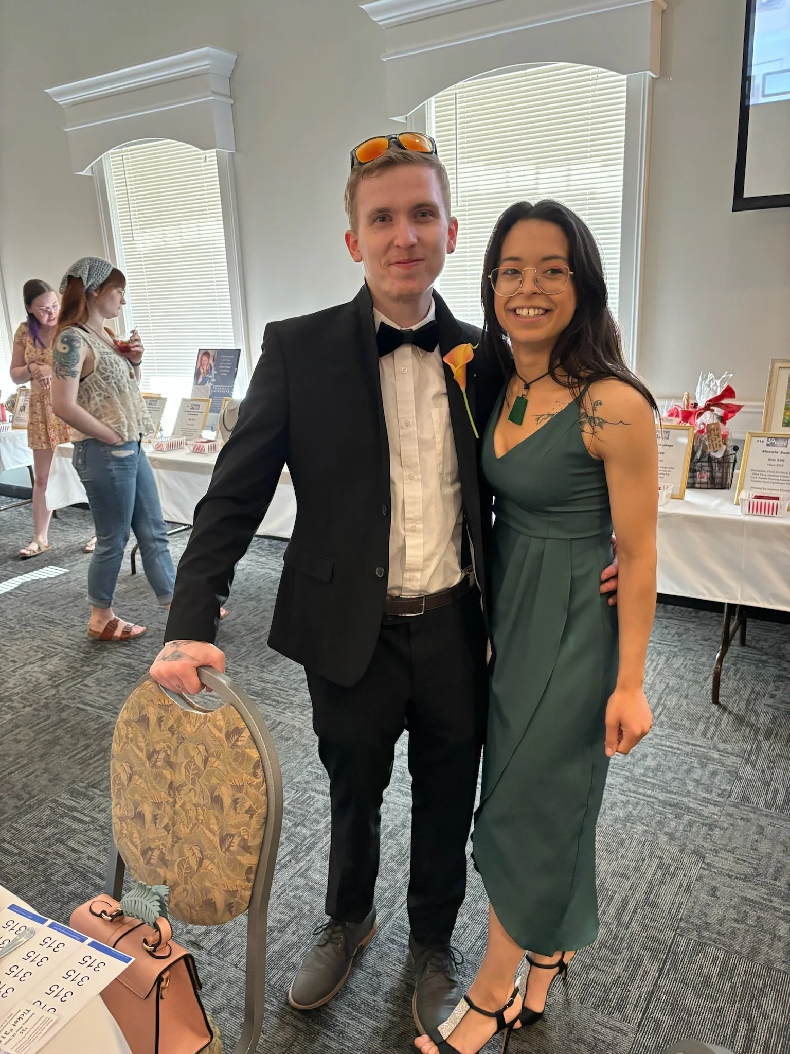 A young man and woman dressed up, smiling and posing together at an indoor event. The man is wearing a tuxedo with a bow tie, sunglasses on his head, and a boutonniere. The woman is wearing a green sleeveless dress and glasses.