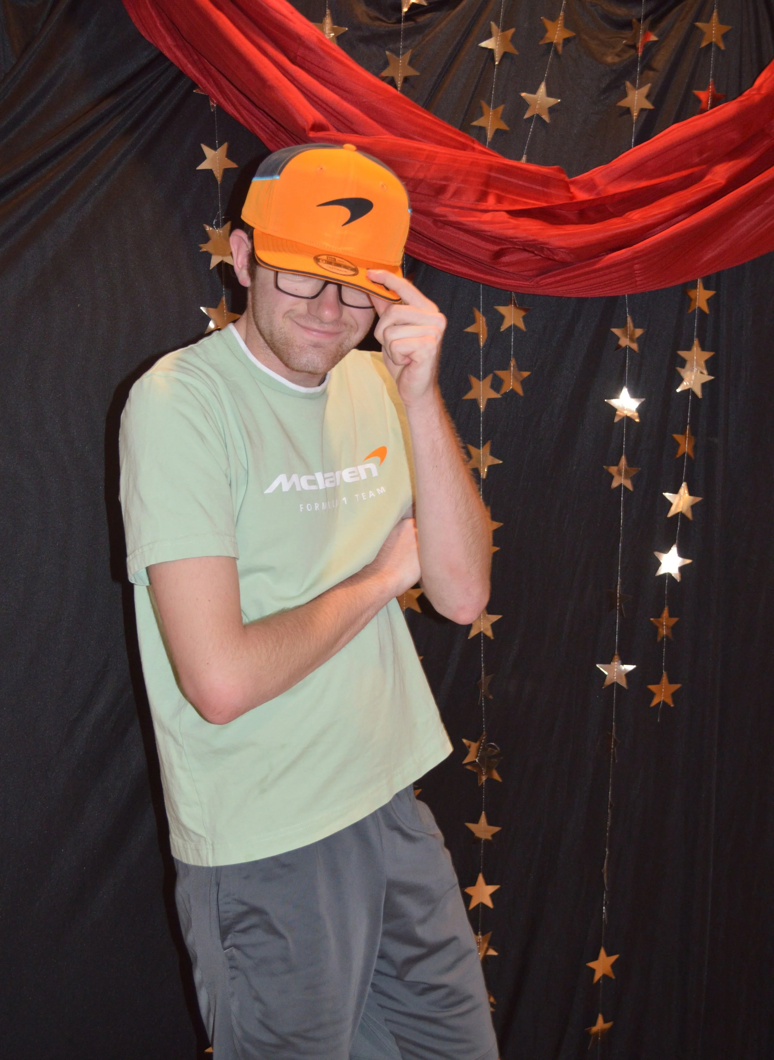 A man wearing glasses, a light green McLaren F1 Team T-shirt, and an orange baseball cap pose with one arm crossed and the other adjusting his cap, standing in front of a backdrop decorated with black fabric, gold star decorations, and red drapery.