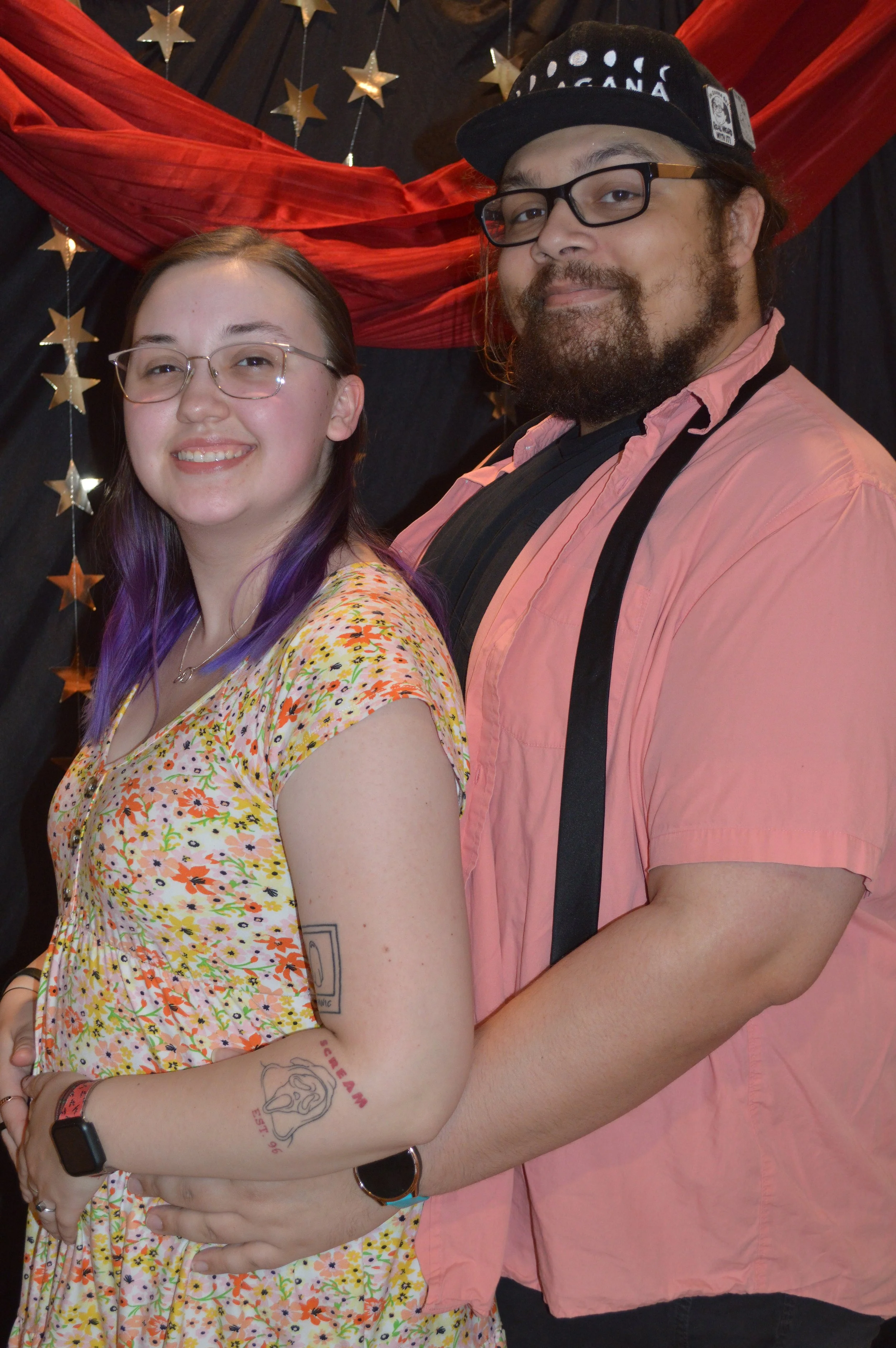 A young woman with glasses and purple-tinted hair, standing in front of a man, both smiling. The woman has tattoos on her arm, one of which reads 'Dream' and features a cartoonish face. They are indoors with a backdrop decorated with stars and red fa