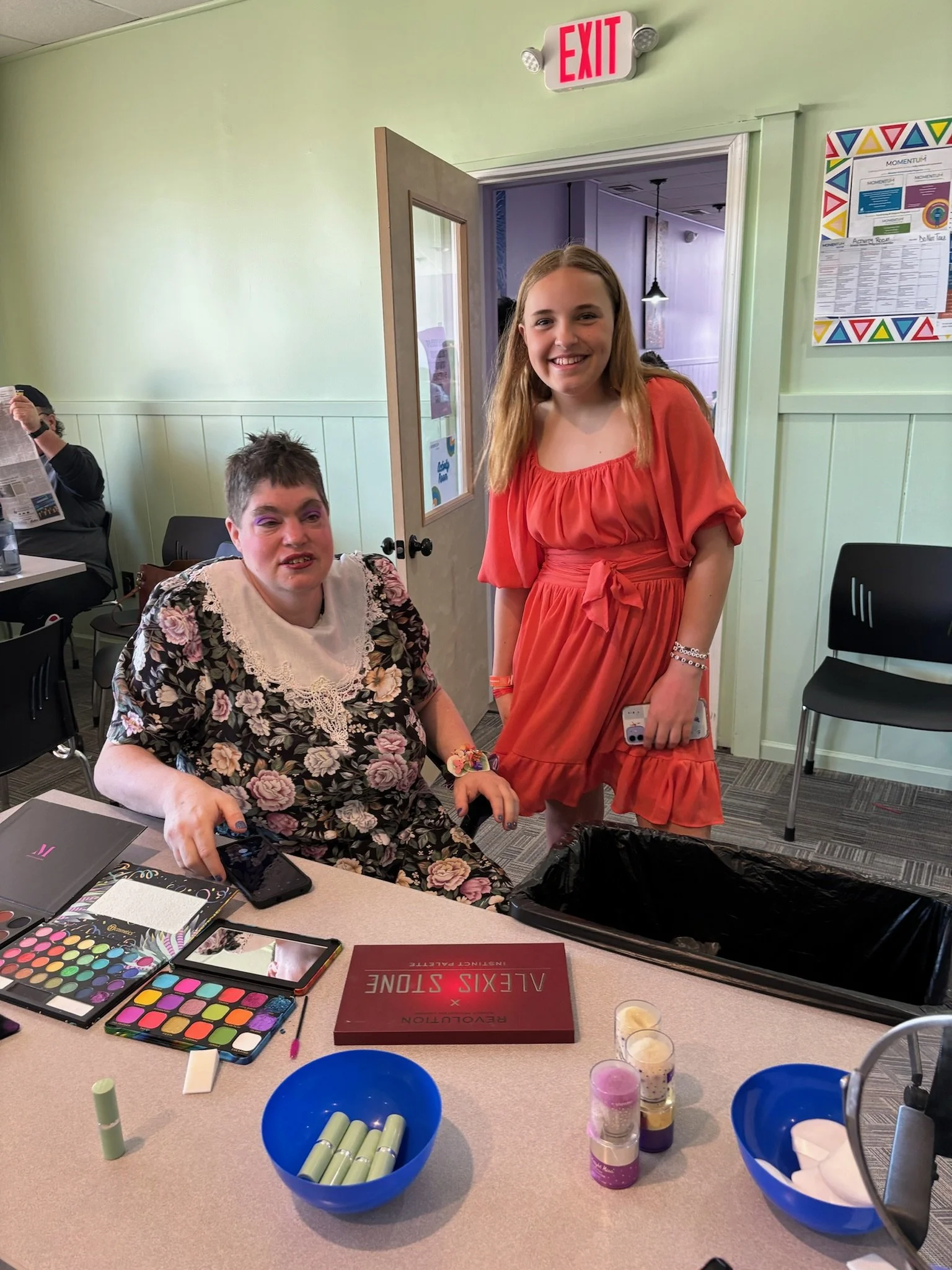 Two young women at a table with makeup products, smiling at the camera. One is seated with a floral dress, and the other is standing in an orange dress. There is an exit sign above the door behind them.