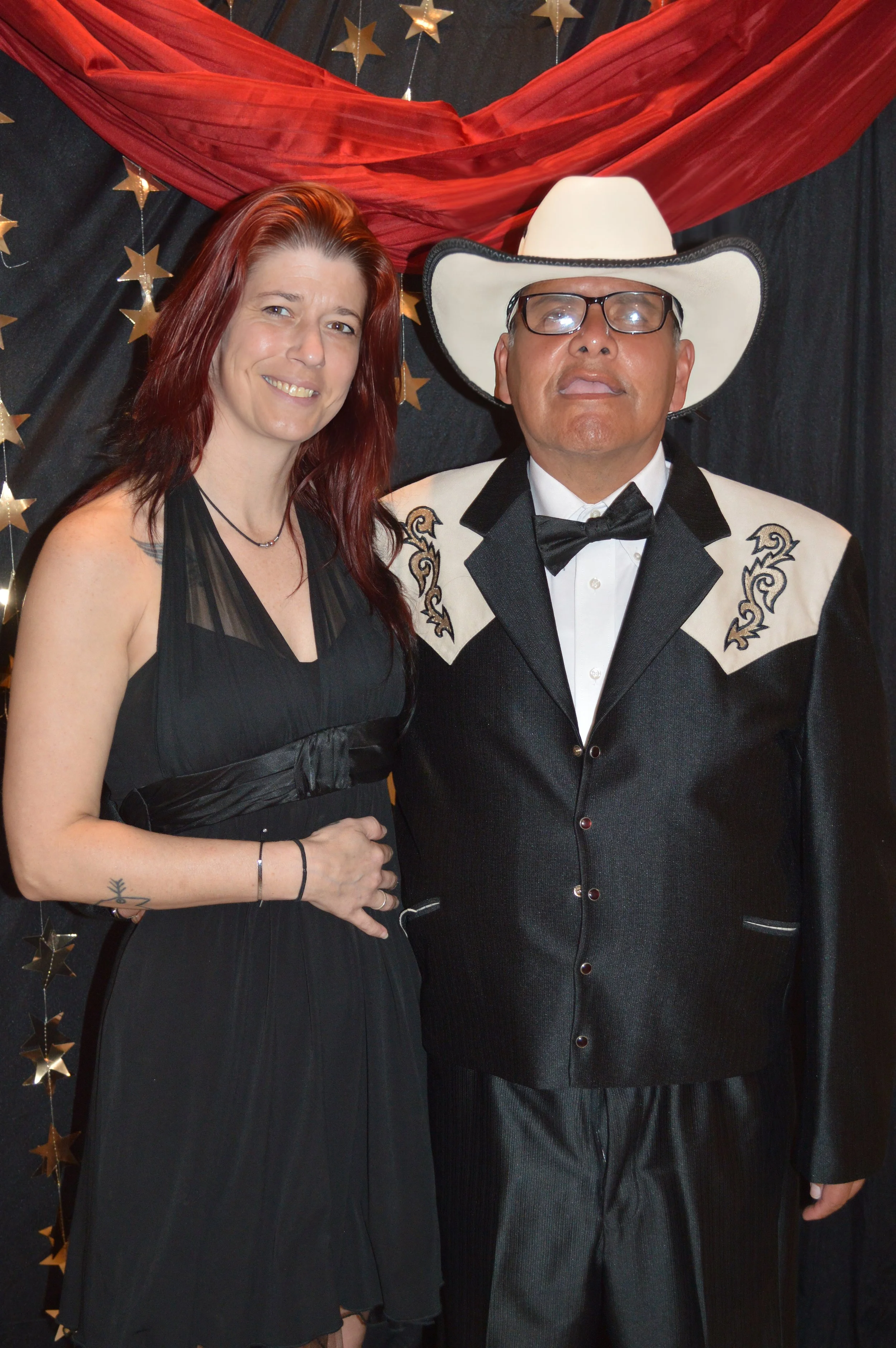 A woman with long red hair and a man dressed as a country singer with a white cowboy hat, glasses, and a black and white western style suit, standing together in front of a red and black decorative backdrop with gold stars.