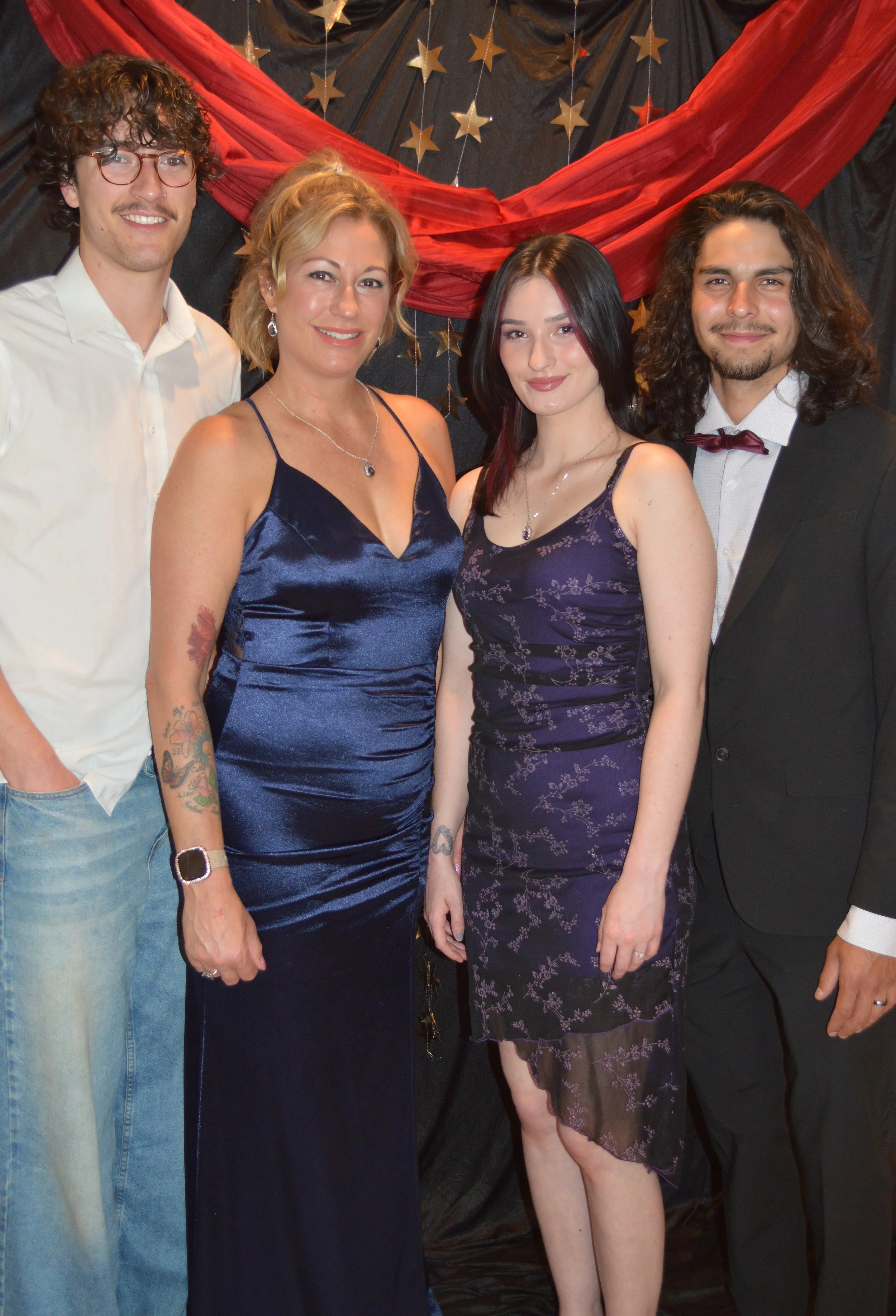 Group of four people at a formal event, posing in front of a decorative backdrop with stars and red fabric. They are dressed in stylish outfits, smiling at the camera.