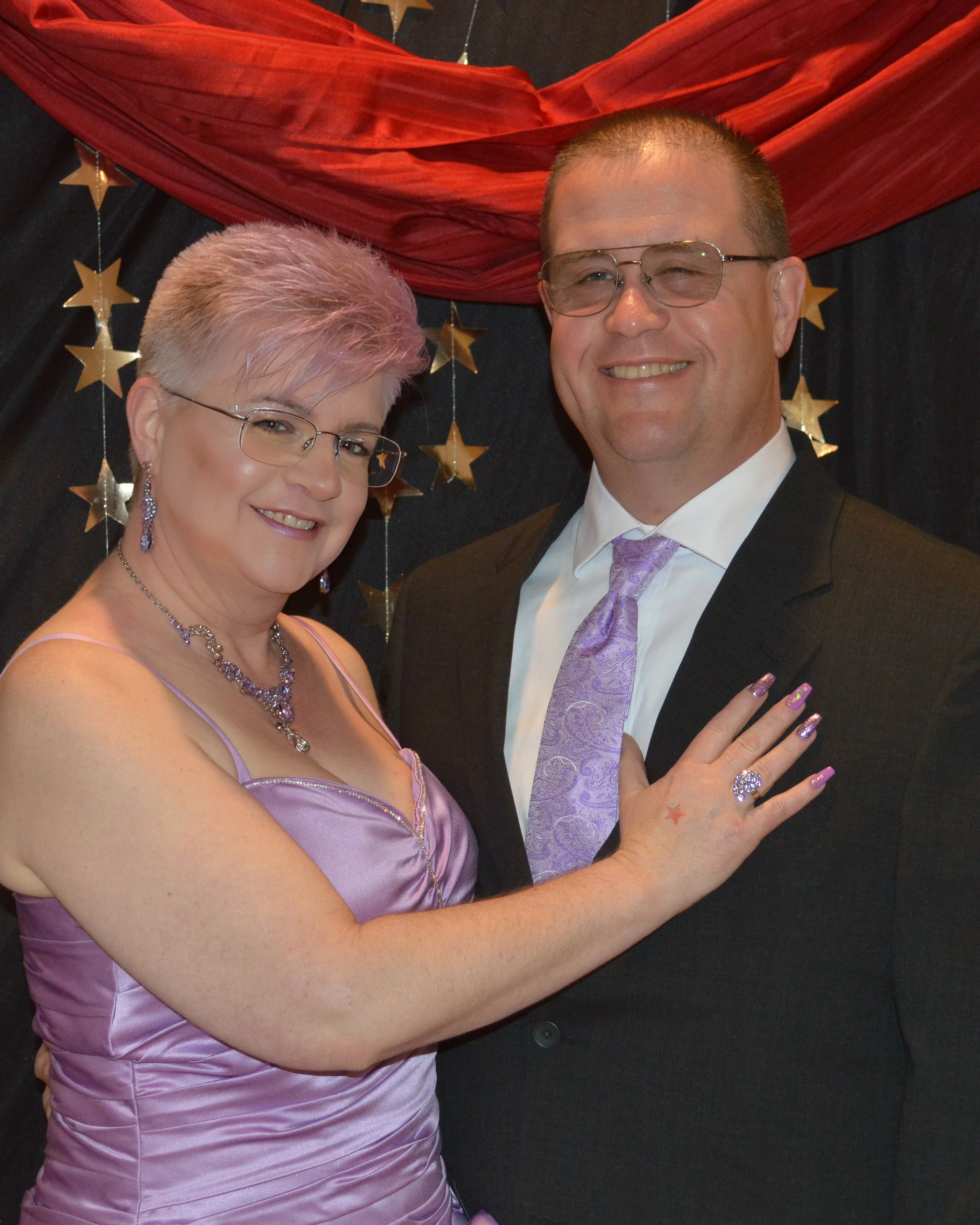 A smiling woman and man dressed in formal attire, standing close together against a star and drape backdrop, with the woman wearing a lavender dress and jewelry, and the man in a black suit with lavender tie.