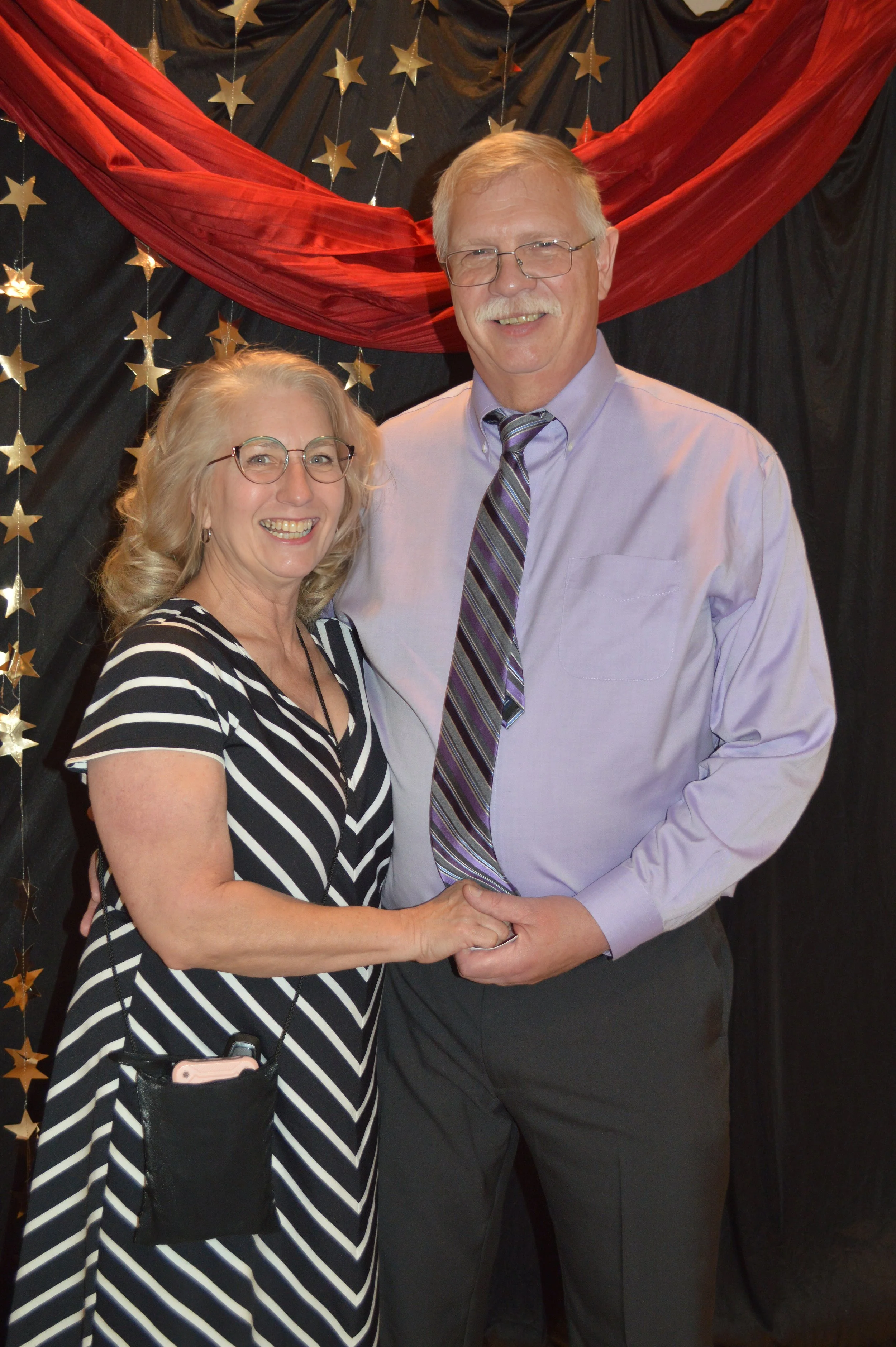 A smiling woman and man holding hands in front of a decorated backdrop with gold stars and red fabric.