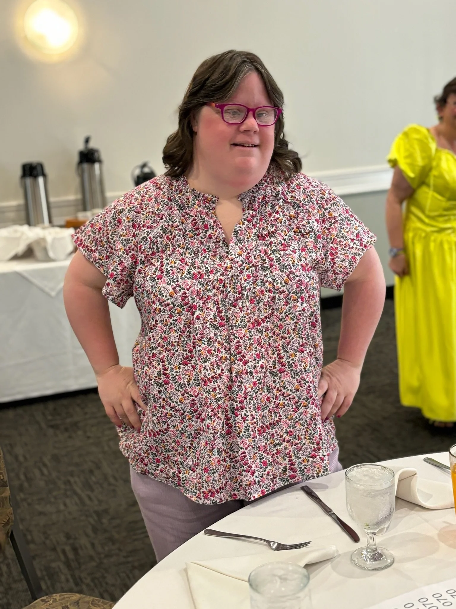 A woman with shoulder-length brown hair, wearing pink glasses and a floral blouse, standing with hands on hips at a table during a social event.