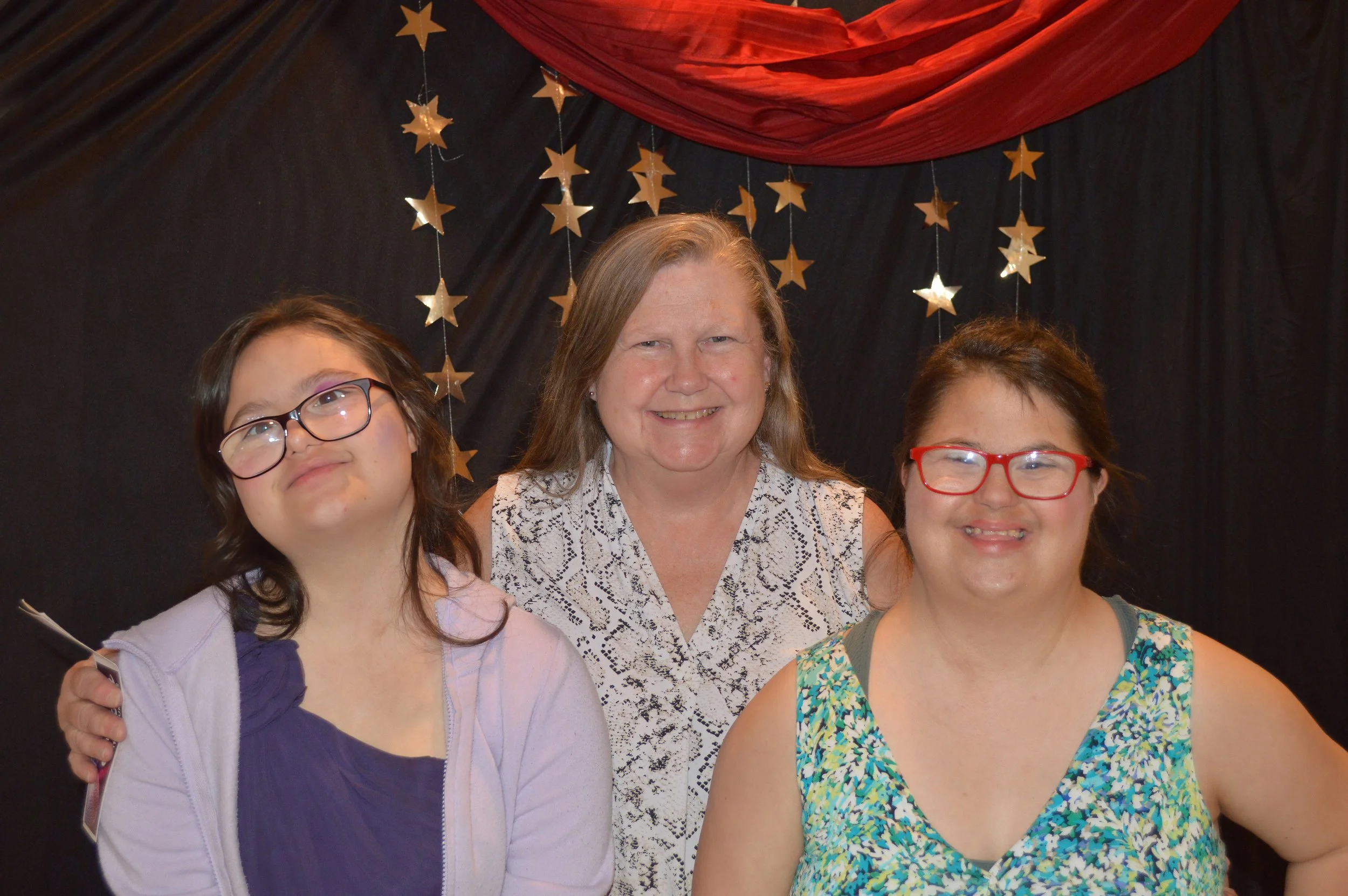 Three women smiling in front of a star and curtain backdrop, two wearing glasses and one without glasses.