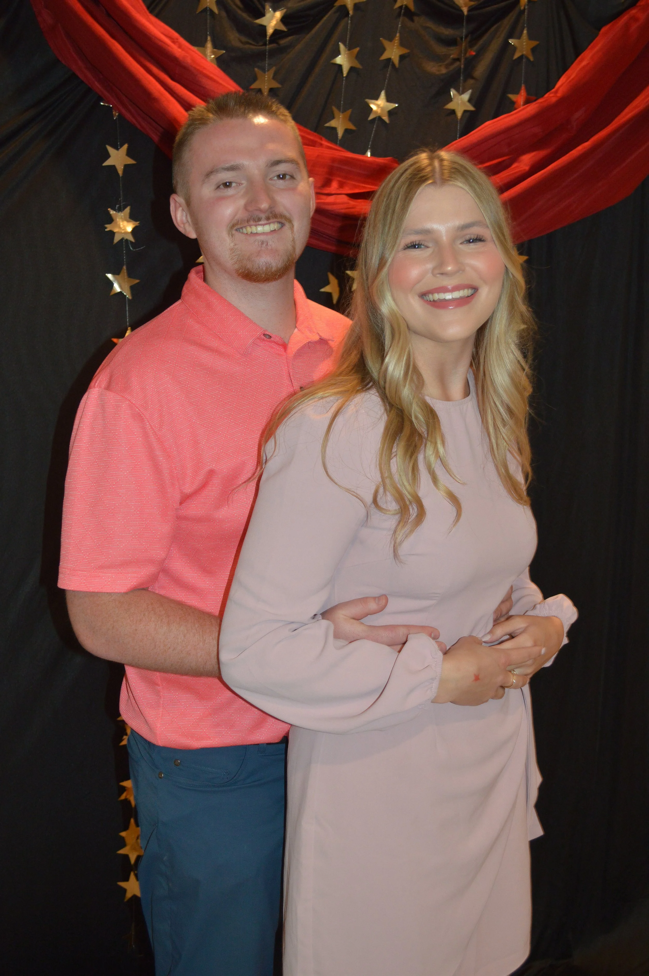 A smiling man and woman pose together, with the man standing behind the woman. The background has a black curtain decorated with gold stars and red fabric. The woman is wearing a light pink dress, and the man is wearing a pink shirt and blue pants.