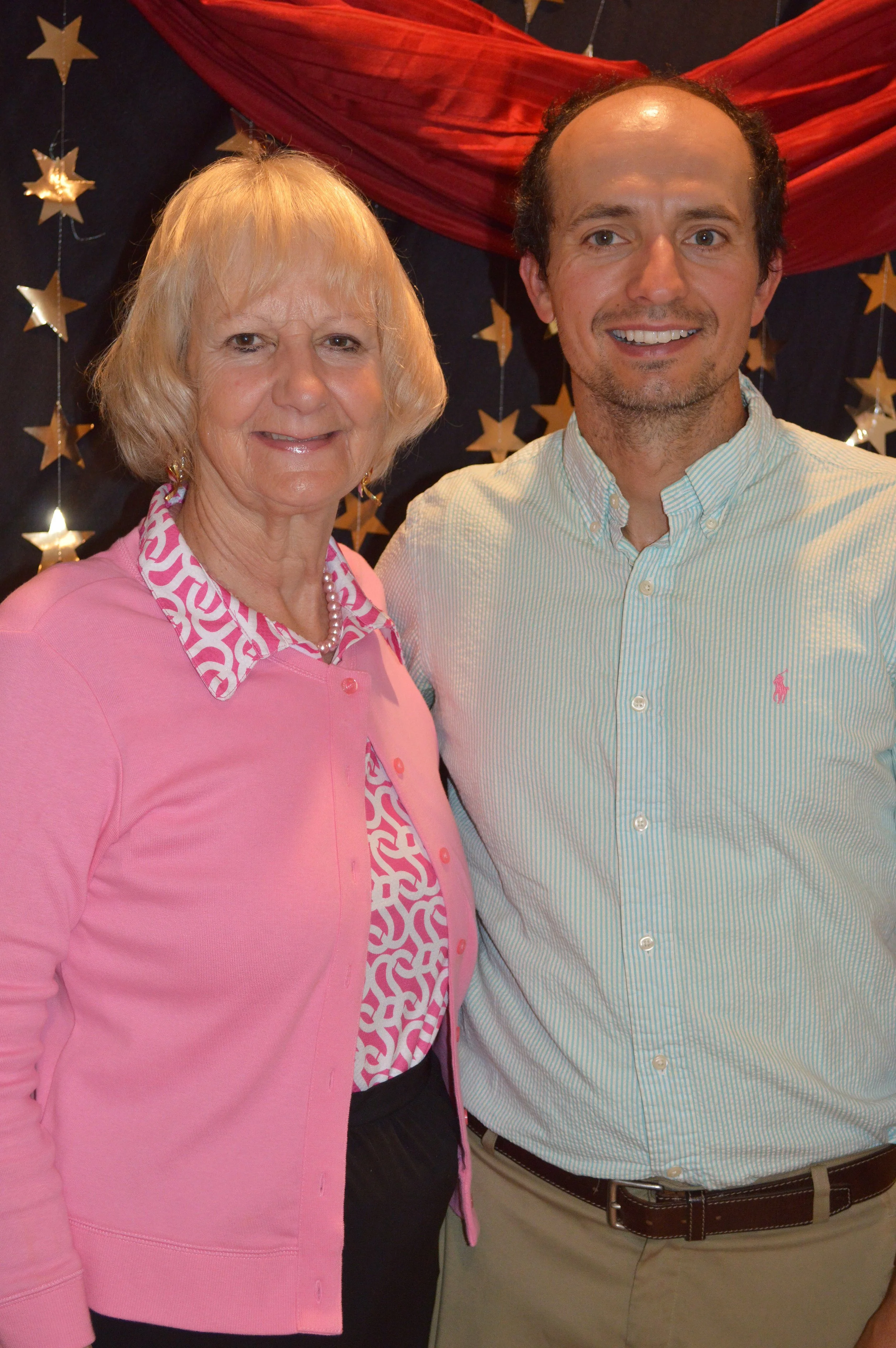 A smiling older woman and a smiling middle-aged man standing close together indoors, with star-shaped decorations and red drapery in the background.