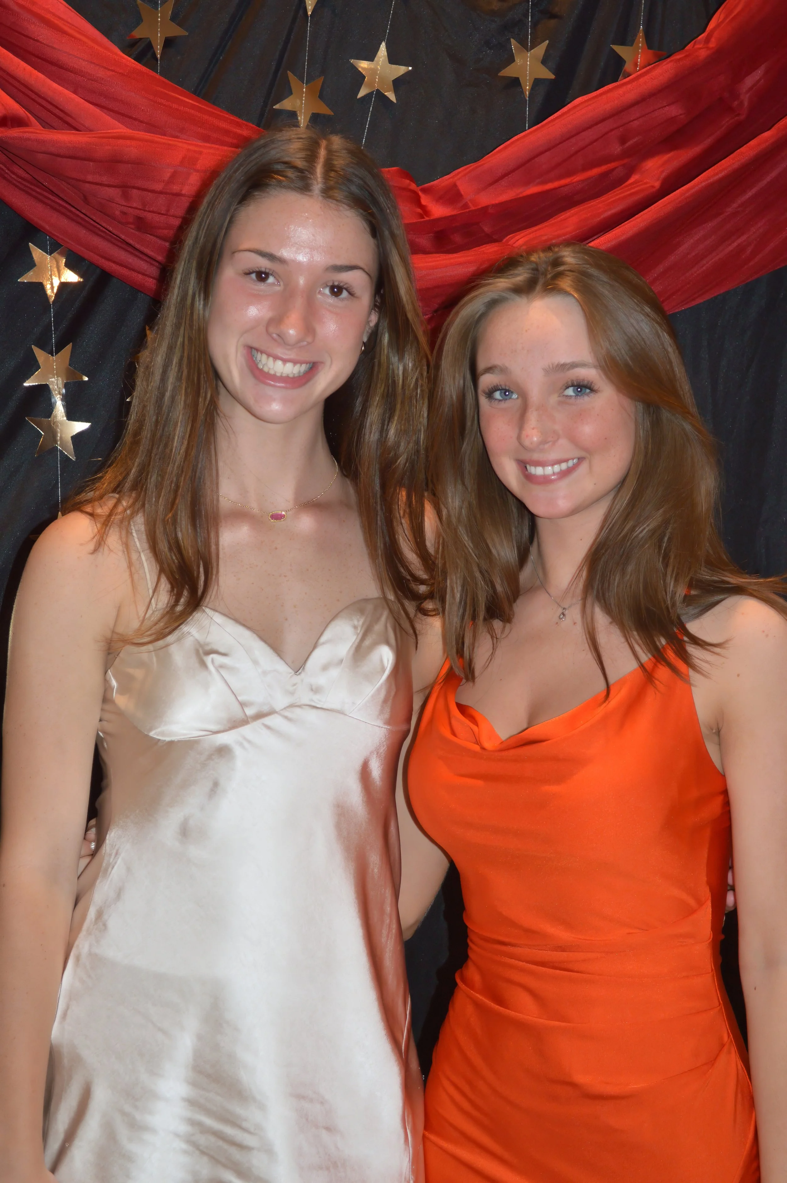 Two young women dressed in formal gowns standing together at a festive event with star decorations and draped fabric in the background.