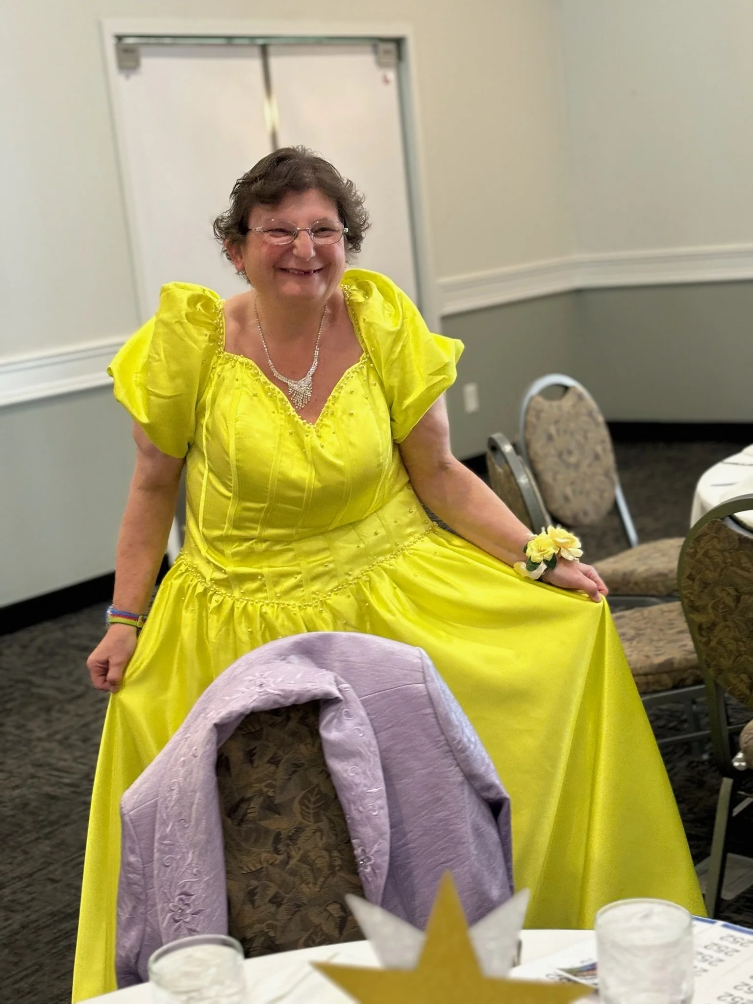 A woman dressed in a bright yellow gown, smiling and holding the edge of her dress, at an indoor event with decorated tables.