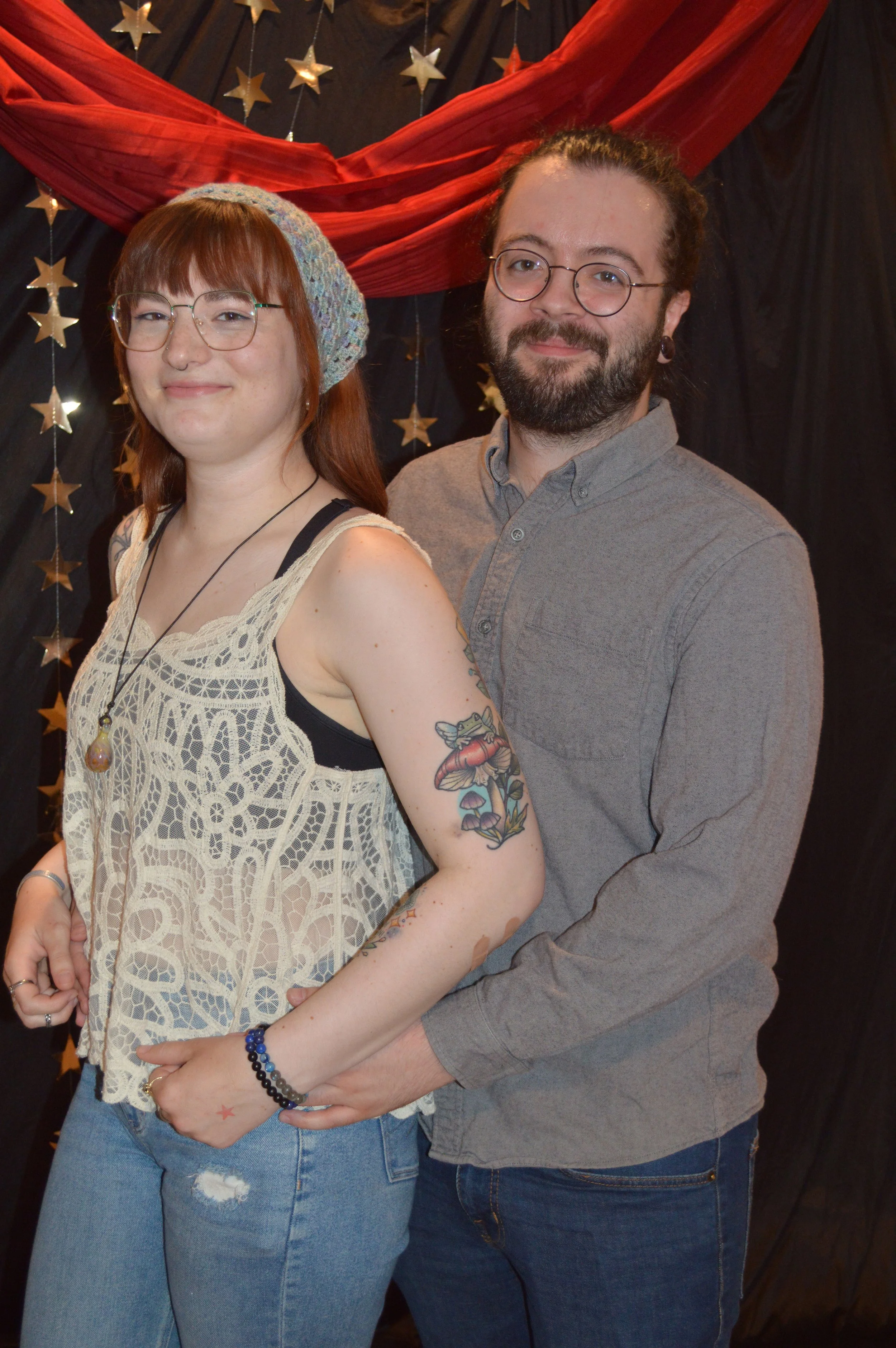 A smiling woman with glasses, a knitted hat, and tattoos on her arm, standing in front of a man with glasses, a beard, and long hair, both posing with a black curtain backdrop decorated with gold stars and a red fabric draped above.
