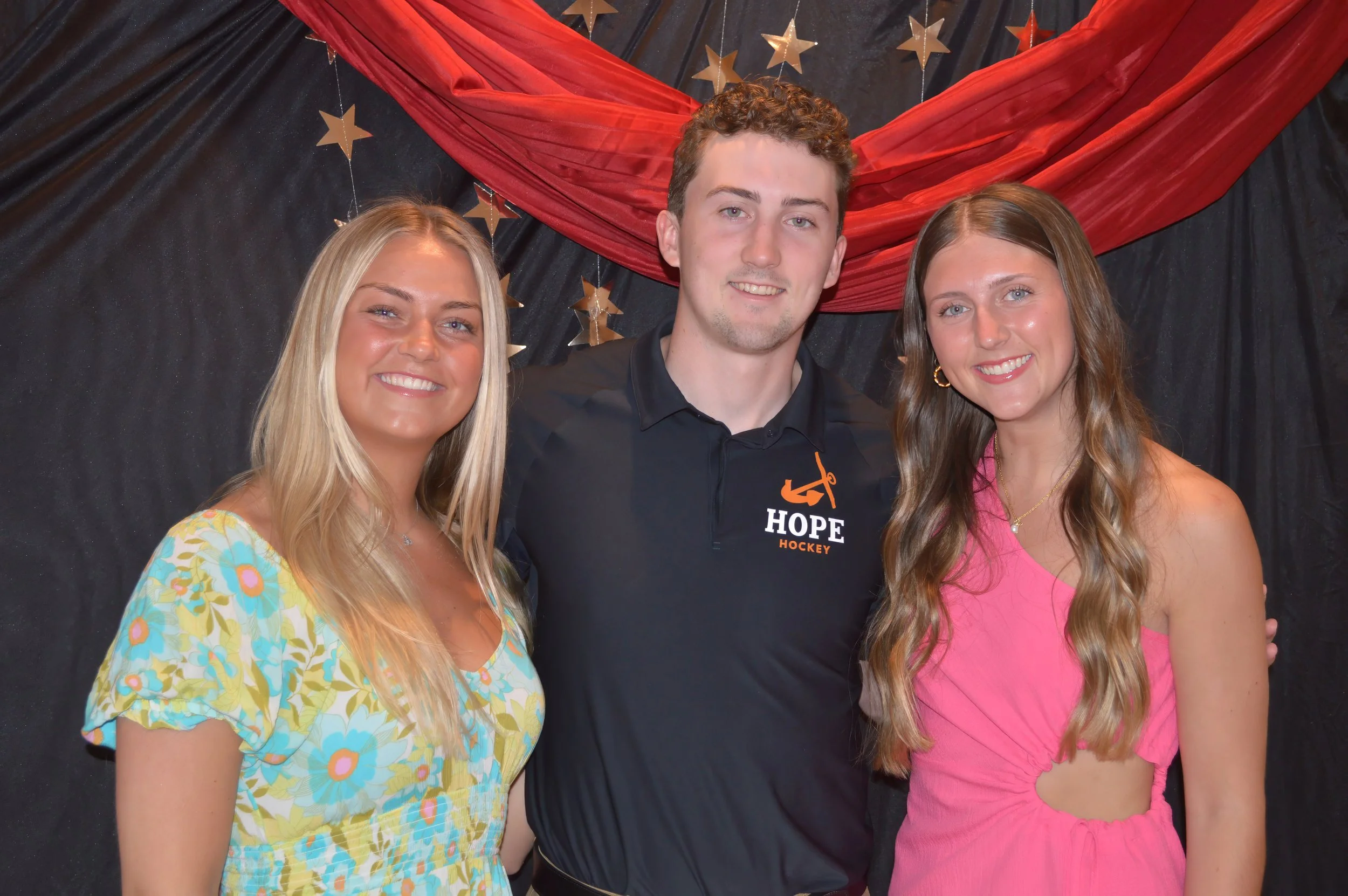 Three young adults, two women and one man, standing together in front of a black backdrop with red fabric and gold star decorations. The man is wearing a black polo shirt with a Hope Hockey logo, and the women are dressed in colorful dresses, one flo