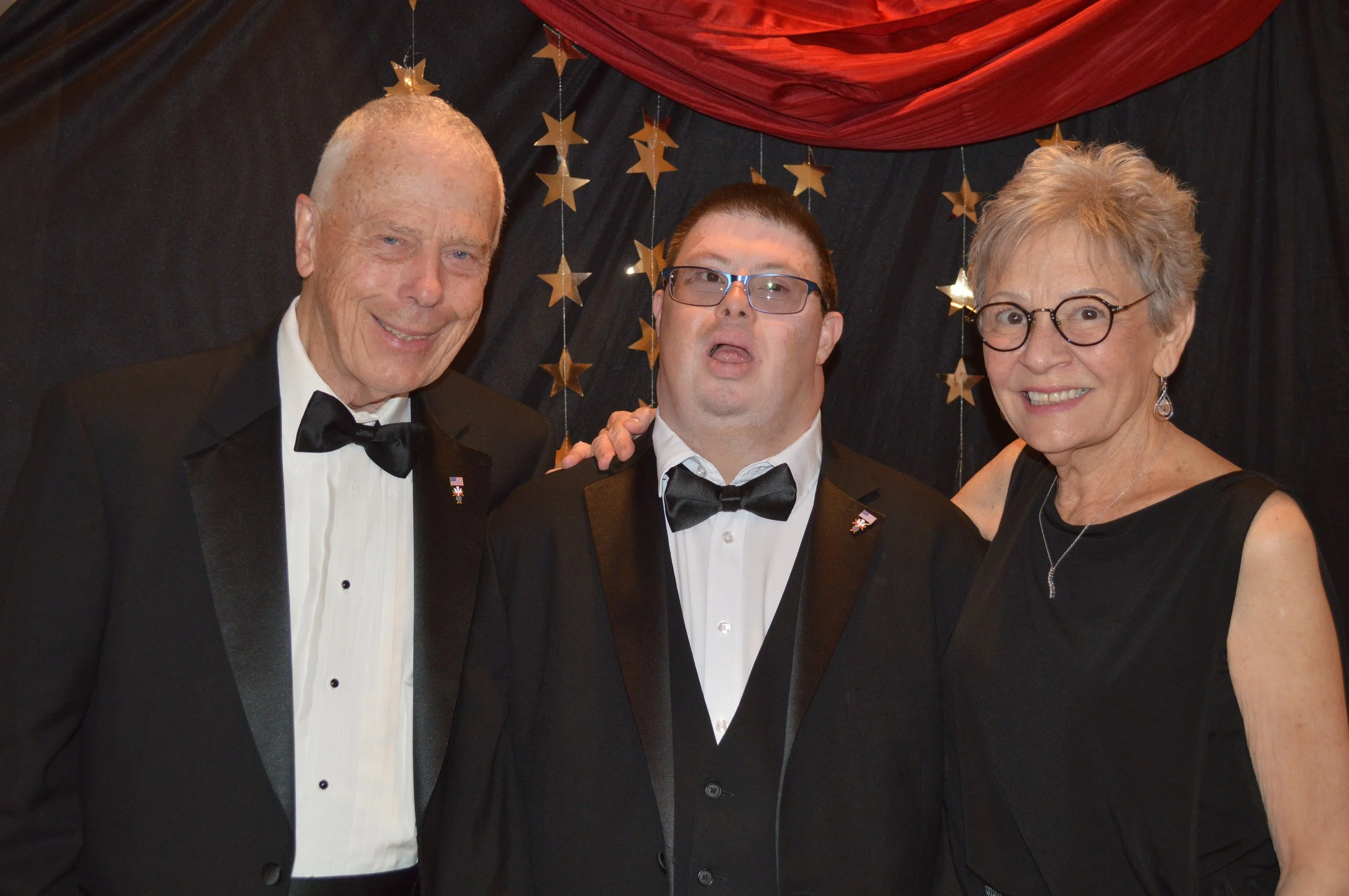 Three people dressed in tuxedos and evening attire standing close together at a formal event, with a backdrop of black fabric decorated with gold star-shaped ornaments and red drape overhead.
