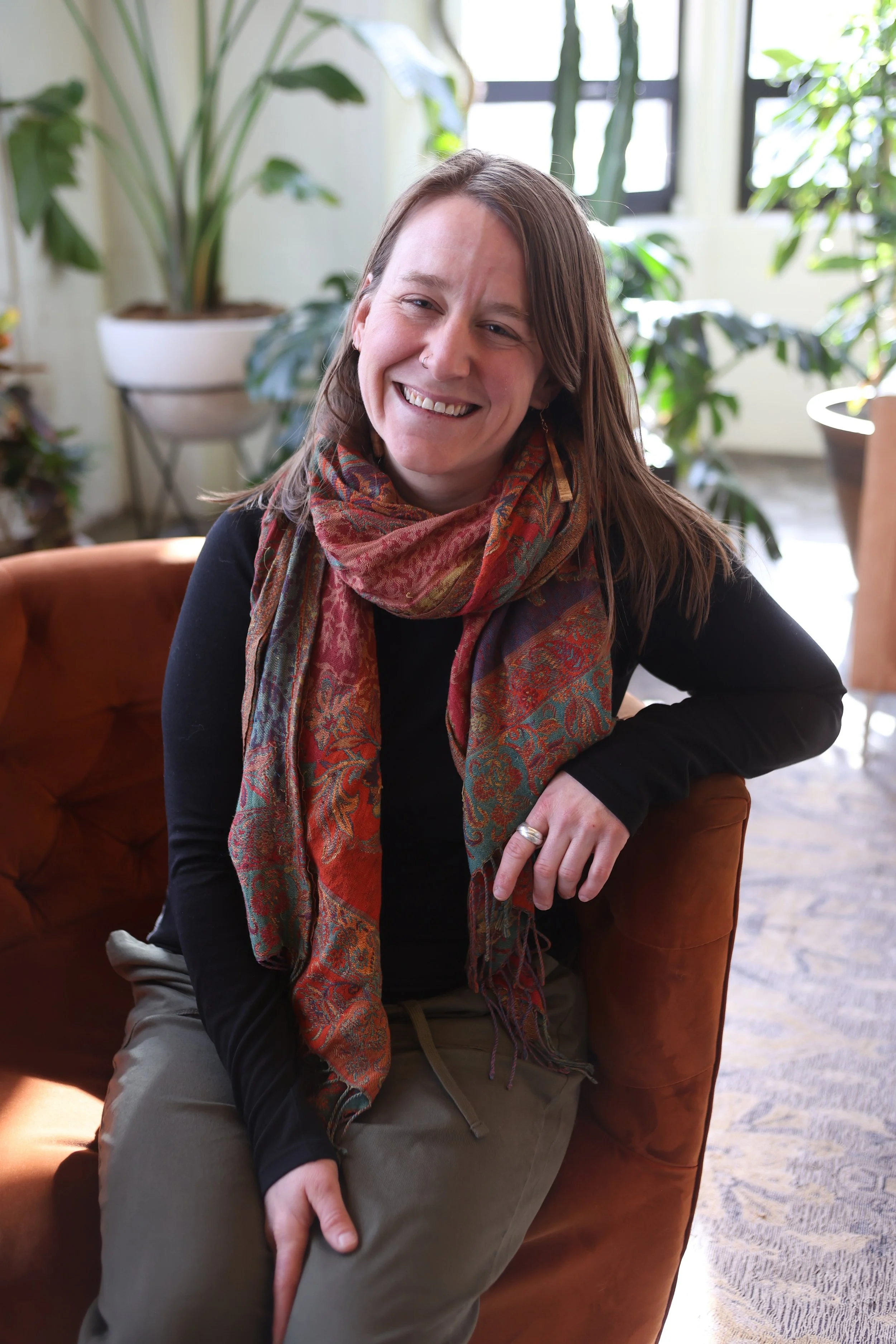 Woman wearing black shirt and colorful scarf sits casually on a sofa and smiles at camera