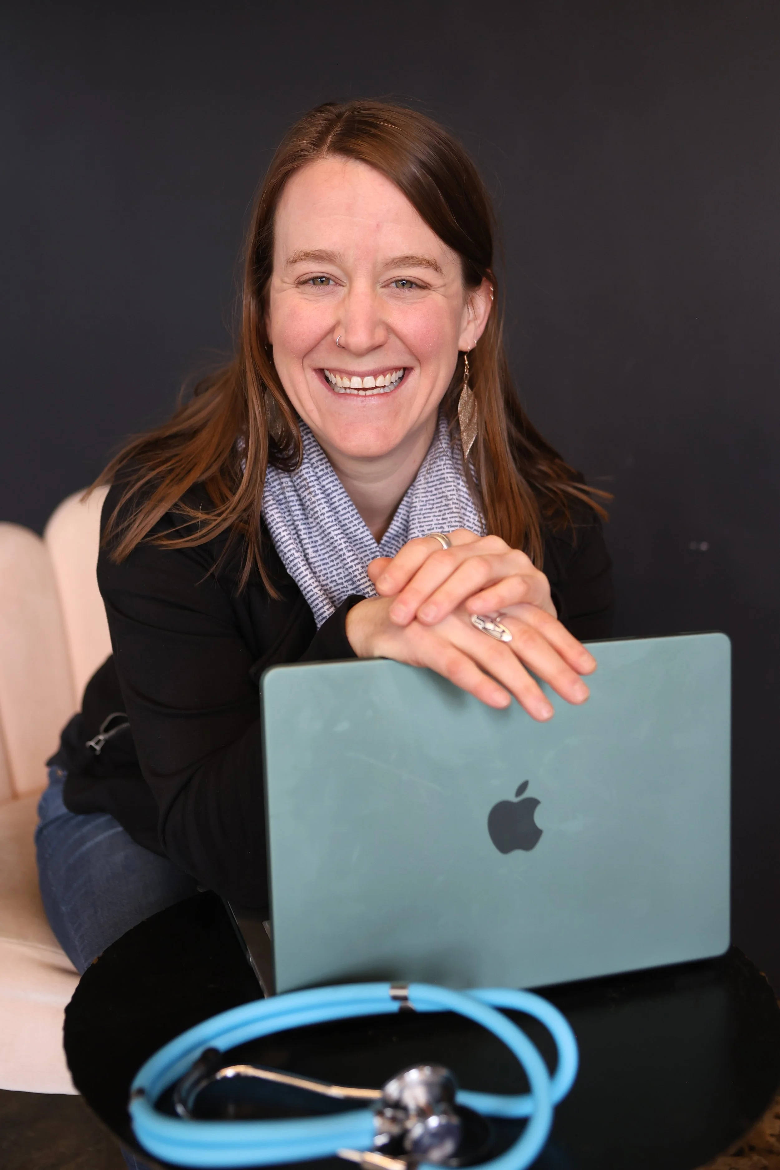 Woman with shoulder length brown hair leans over a sage green laptop and smiles at camera