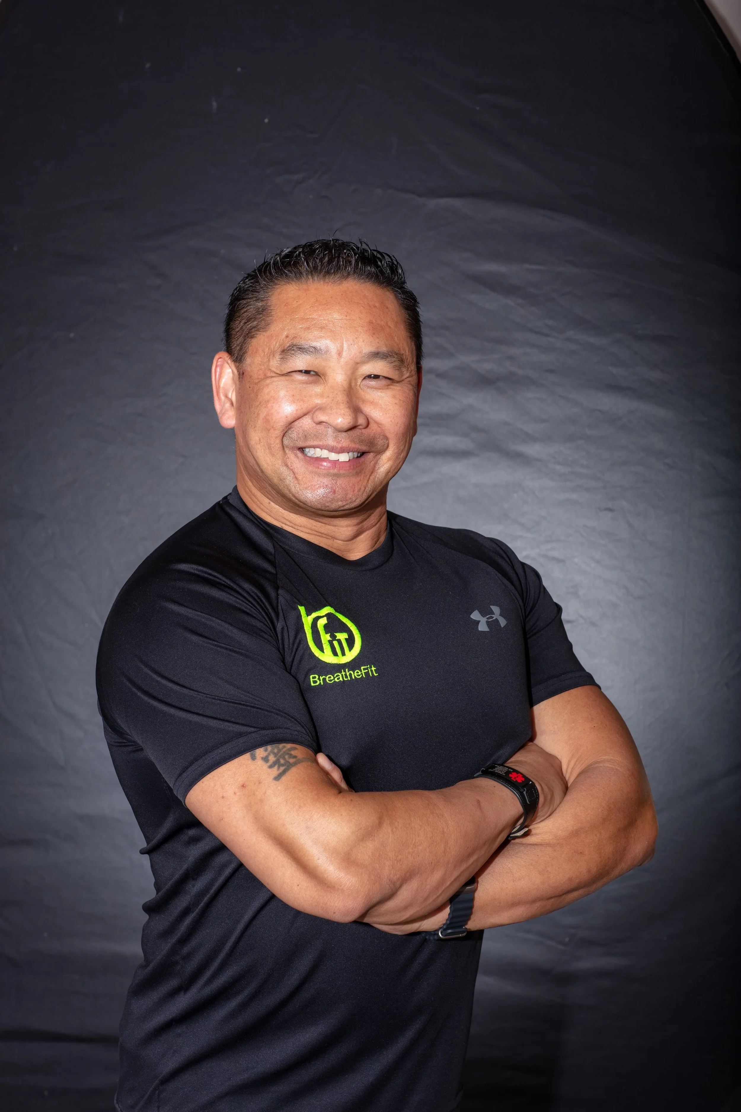 A smiling man with crossed arms, wearing a black athletic shirt with 'BreatheFit' logo, against a grey background.