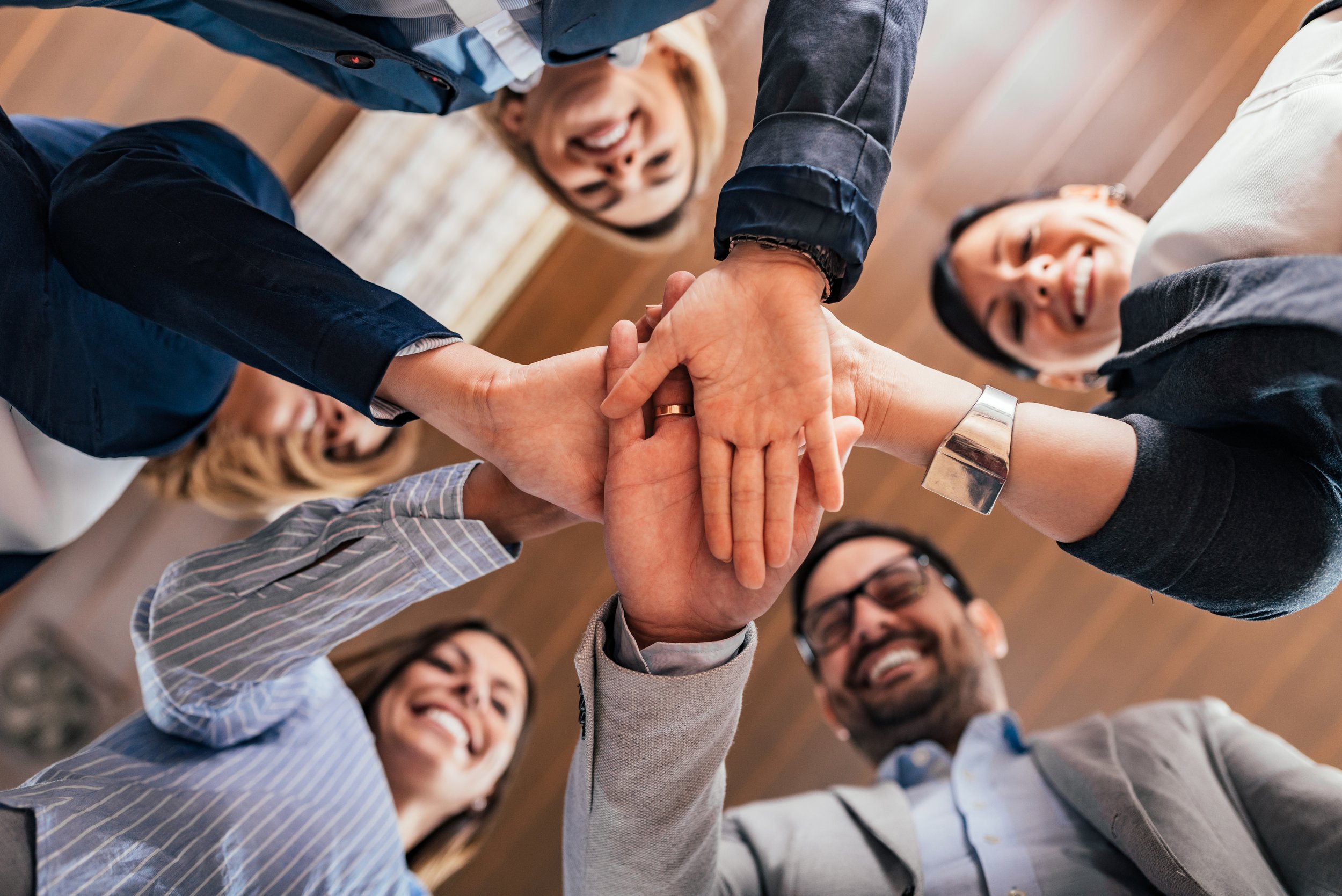 A circle of professionals are facing each other and smiling and putting their hands together in the middle of the circle to demonstrate team work.