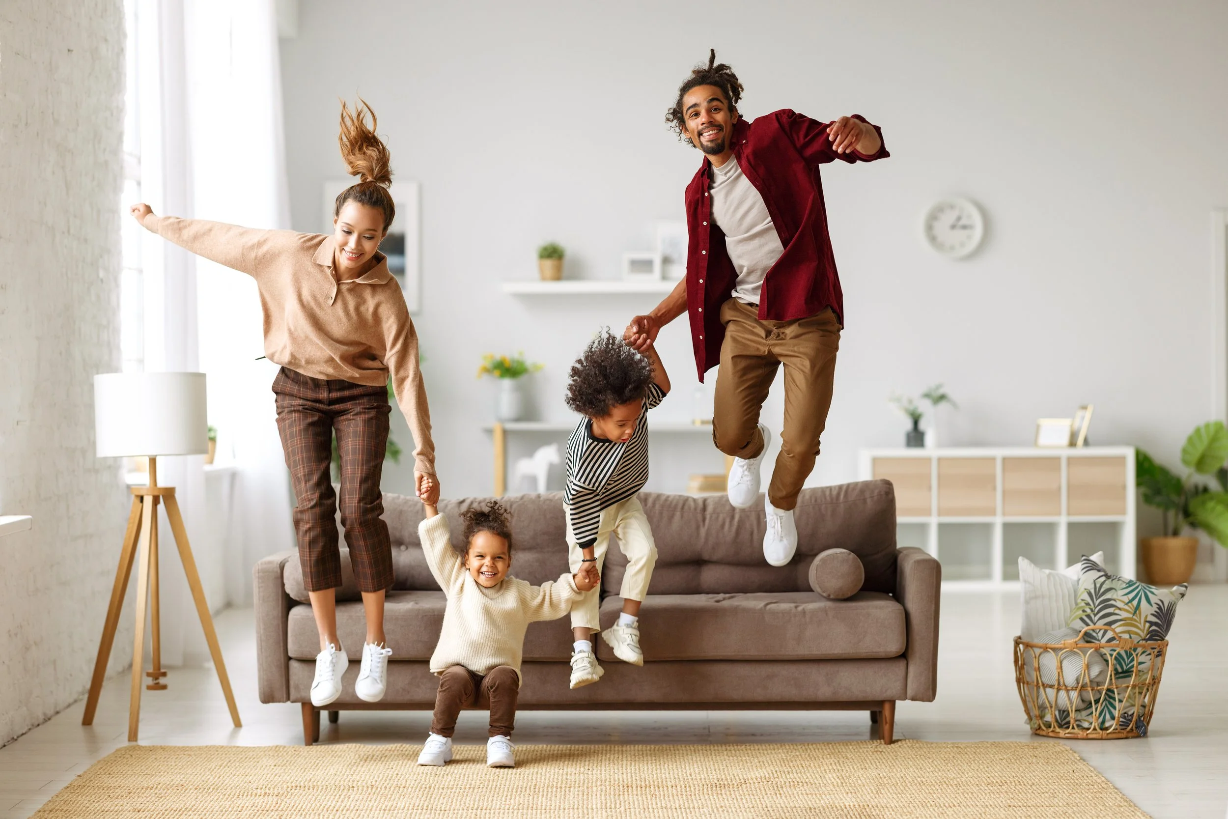 Two parents with toddler aged children jump off a couch together in a living room while holding hands and smiling.