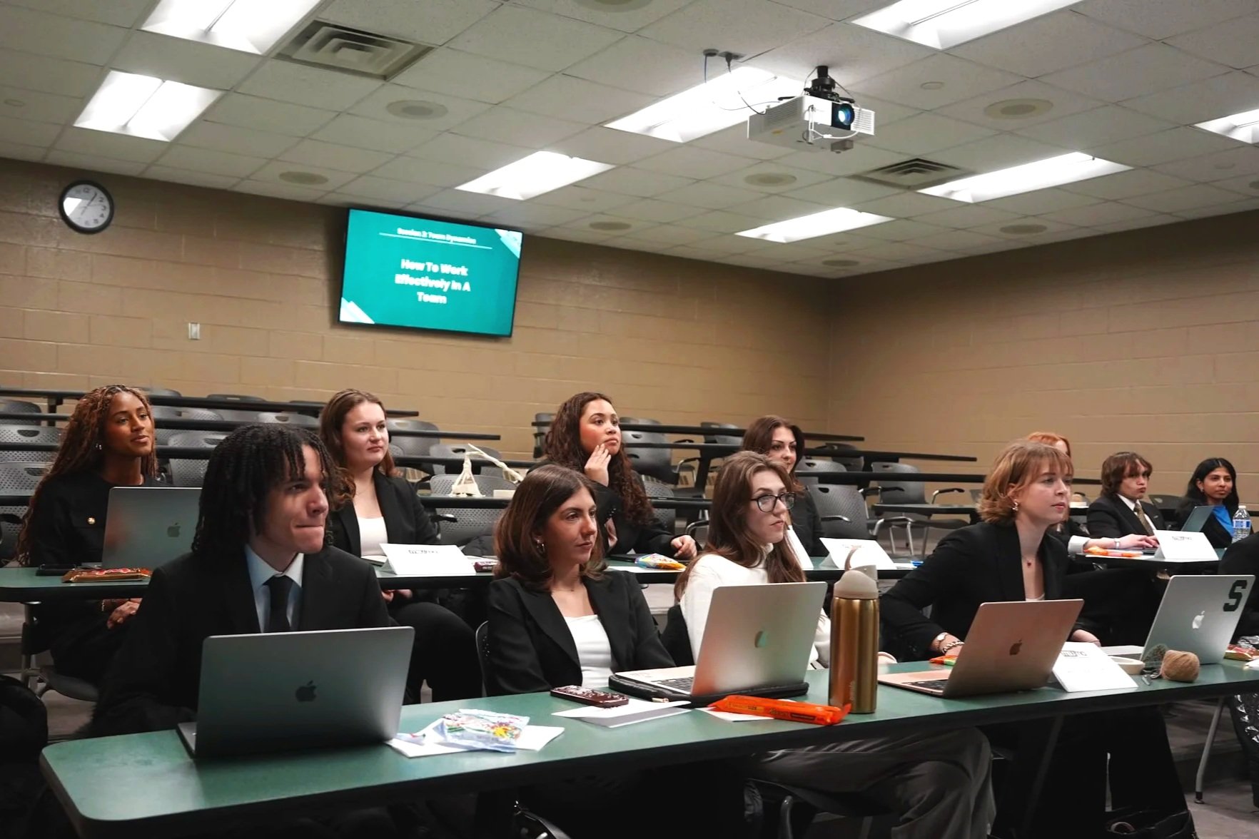 Four women are giving a presentation on stage in front of an audience. Behind them is a large screen showing a SWOT analysis with icons and text about strengths, opportunities, weaknesses, and threats. The background of the stage has a banner with the logo and name of the institution, Broad College of Business.