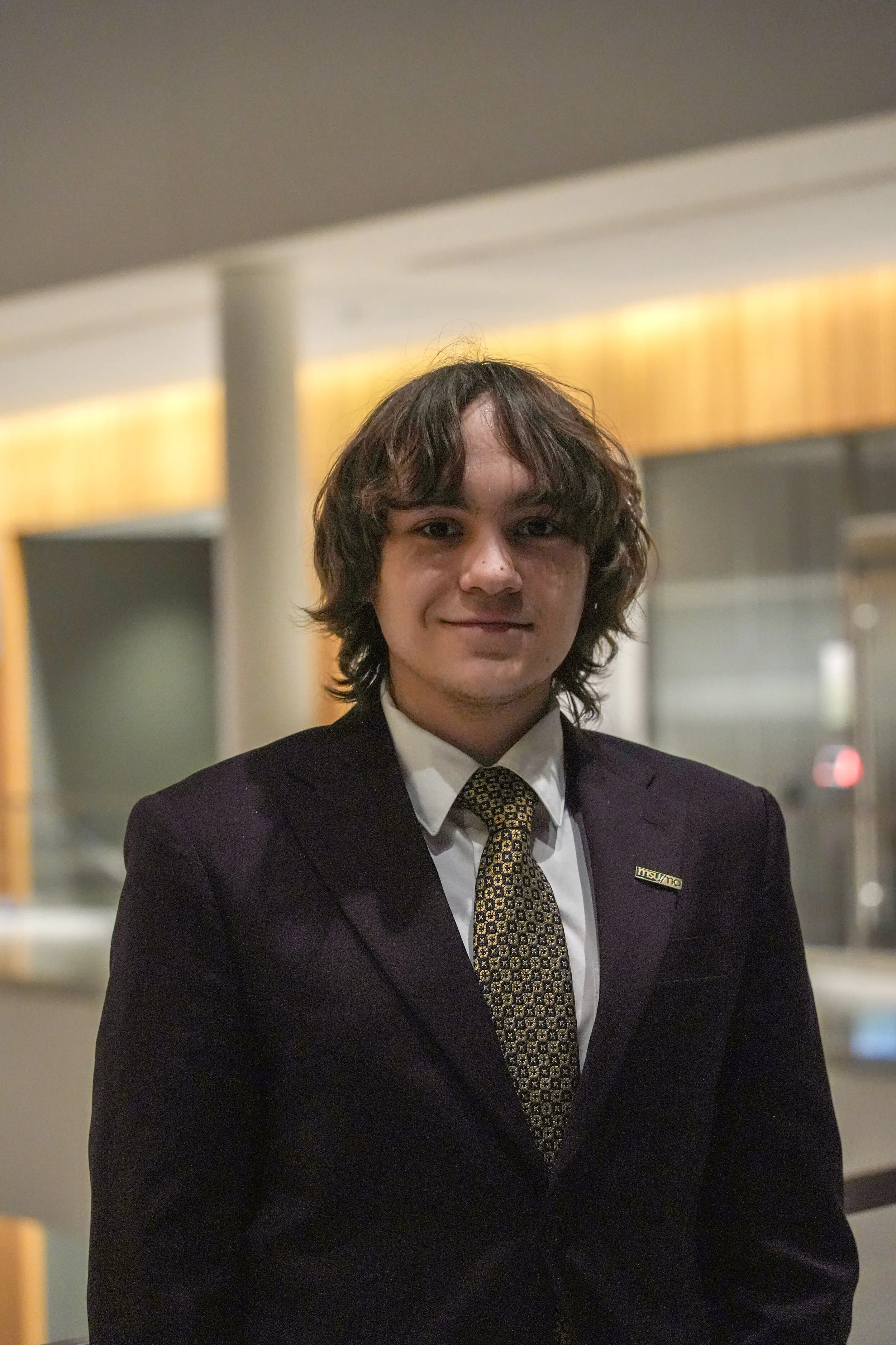 Michigan State University Spartan helmet logo with MSUMA text, in front of a modern building with glass windows and trees.