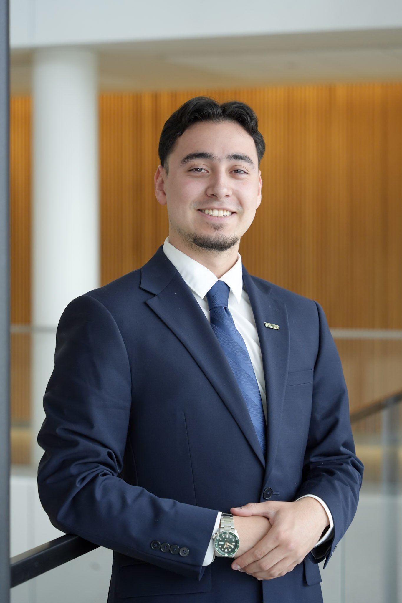 Professional portrait of a young man with dark curly hair, light skin, wearing a navy suit, white shirt, and navy tie, smiling at the camera against a gray background.