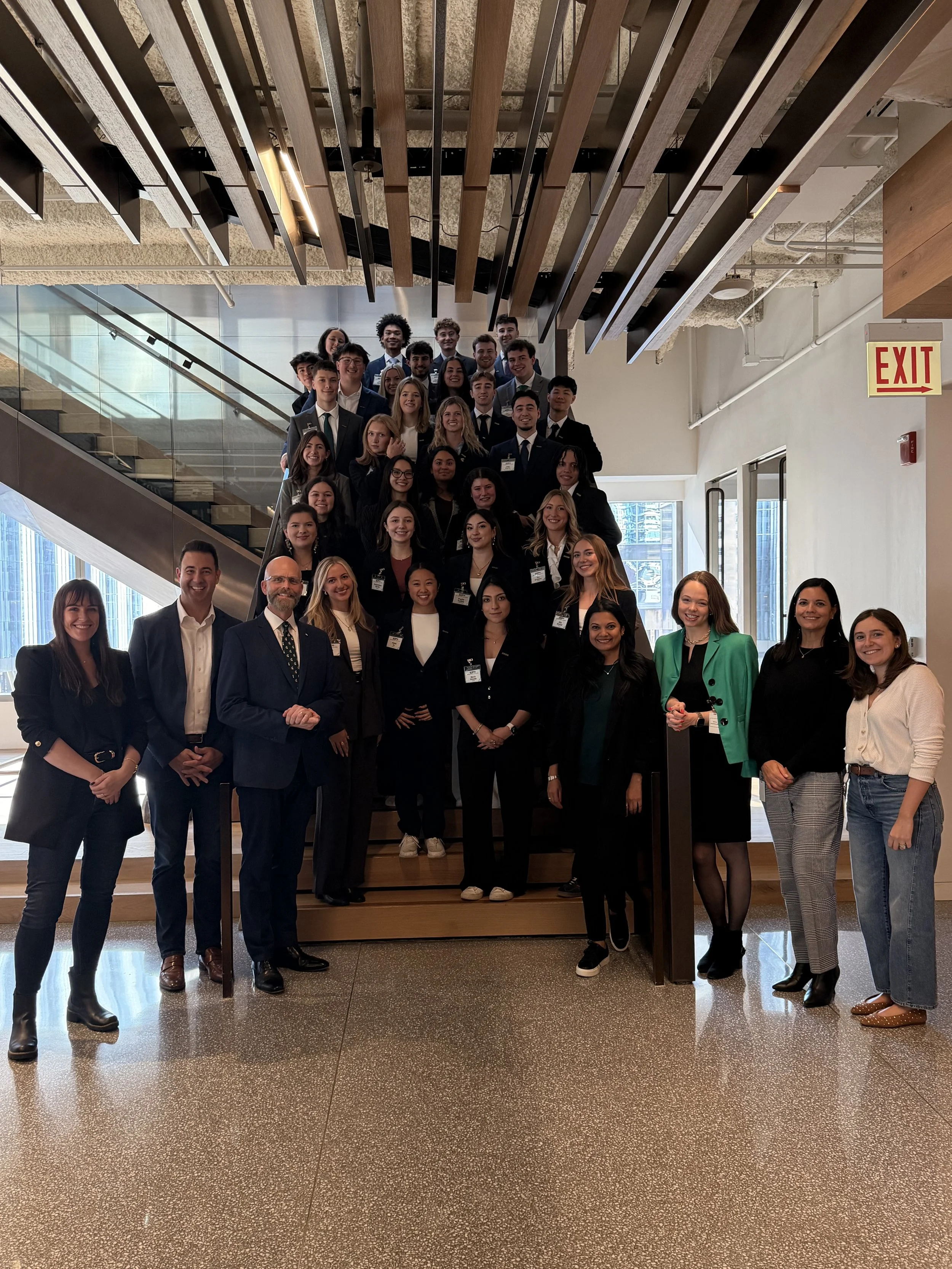 Group of professionally dressed young adults and older professionals posing on a staircase in a modern building, with some standing on the stairs and others at the bottom, smiling at the camera.