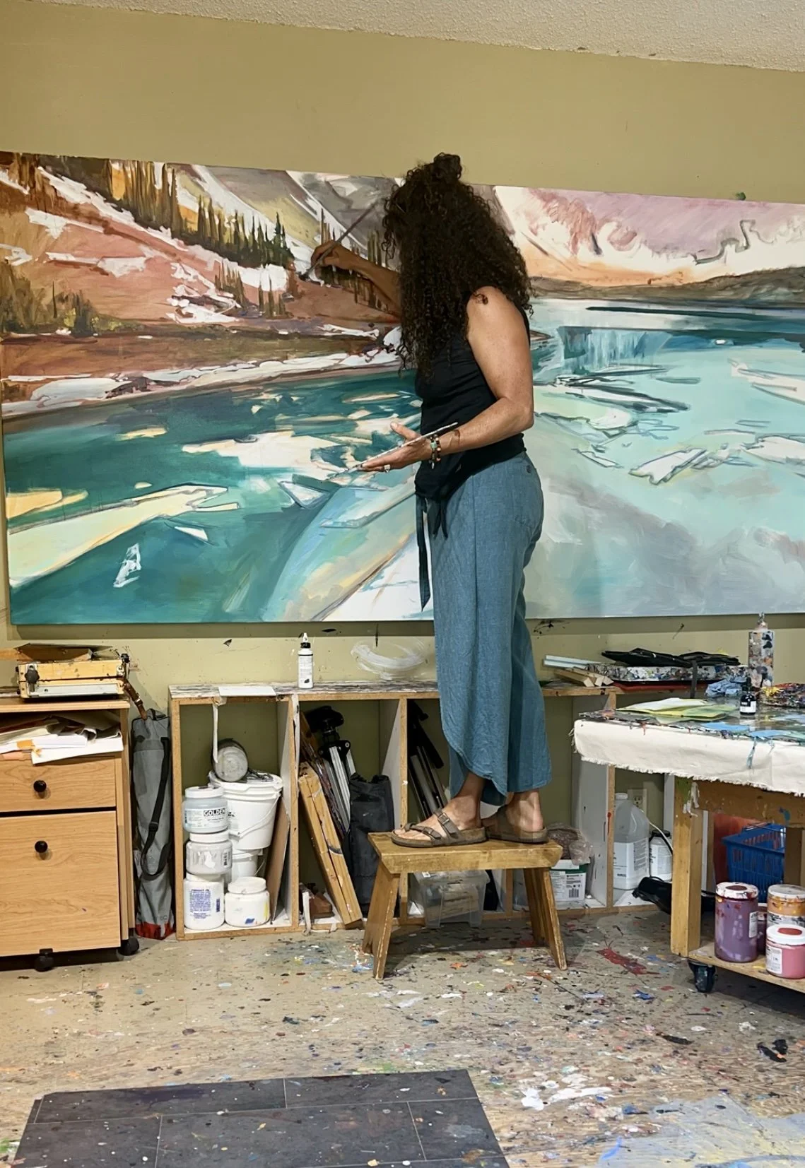 An artist with long curly hair painting a landscape on a large canvas in her studio, standing on a small wooden step stool.