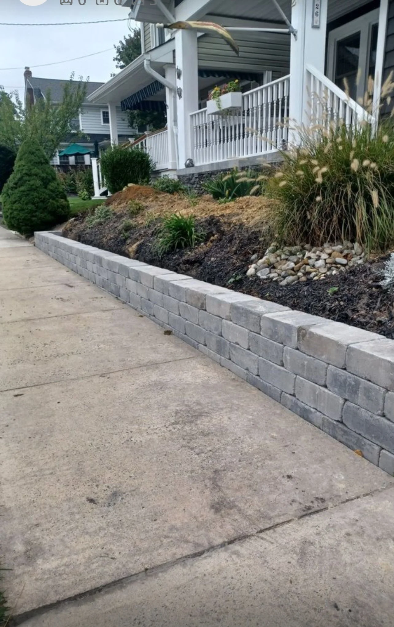 Sidewalk next to a garden with a low stone retaining wall, grass, shrubs, and a house with a porch.