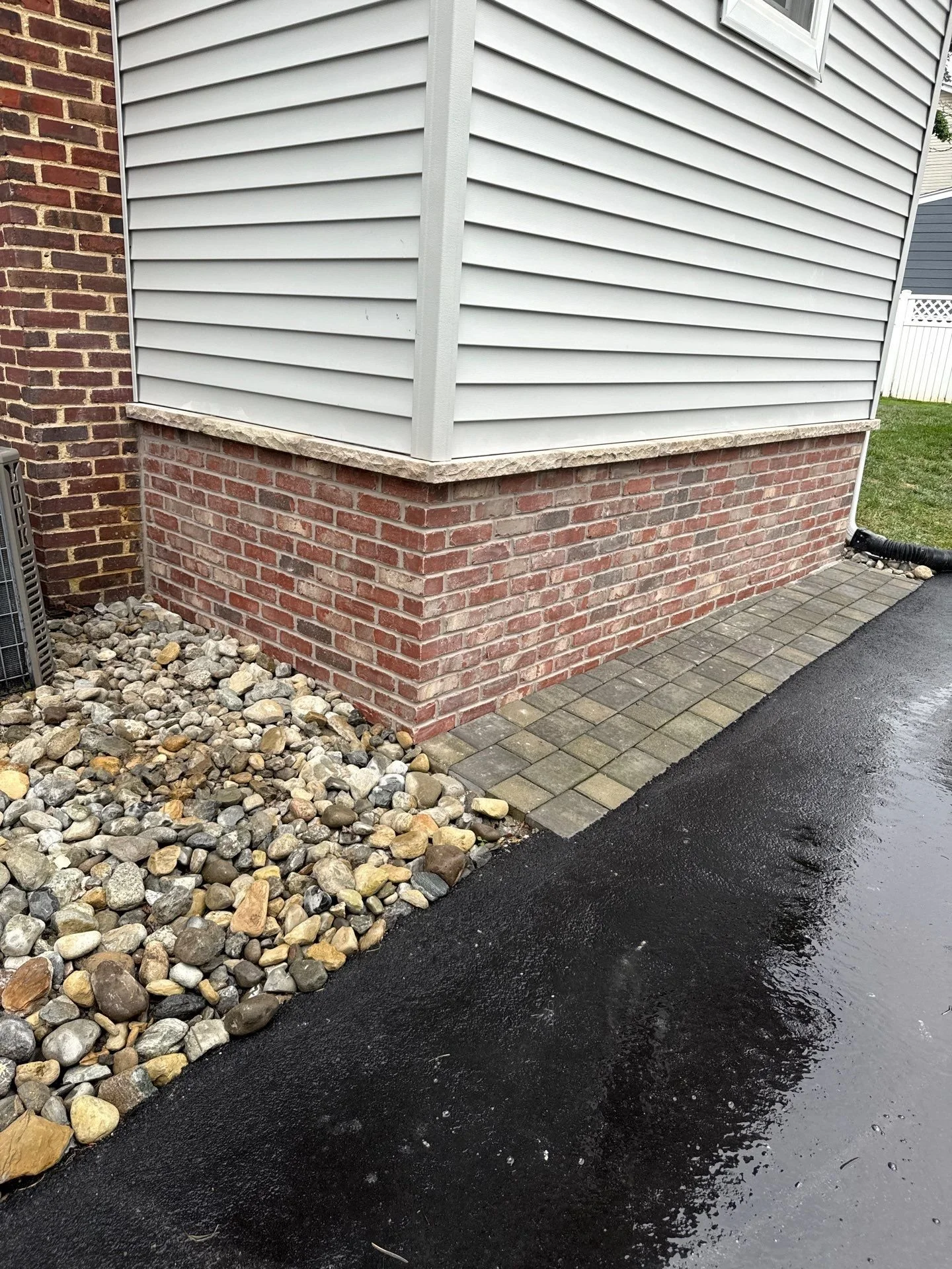 Brick veneer base wrapping a home's corner in Little Silver, NJ featuring a natural stone cap, a paver border, and a river-rock drainage bed along the driveway.