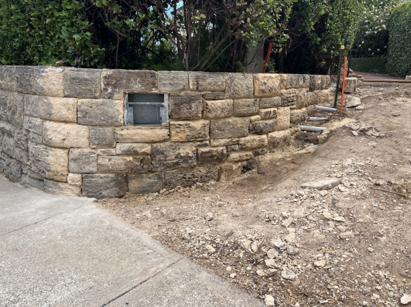 Stone retaining wall with a small window, adjacent to a dirt pathway and stairway under construction, surrounded by greenery.