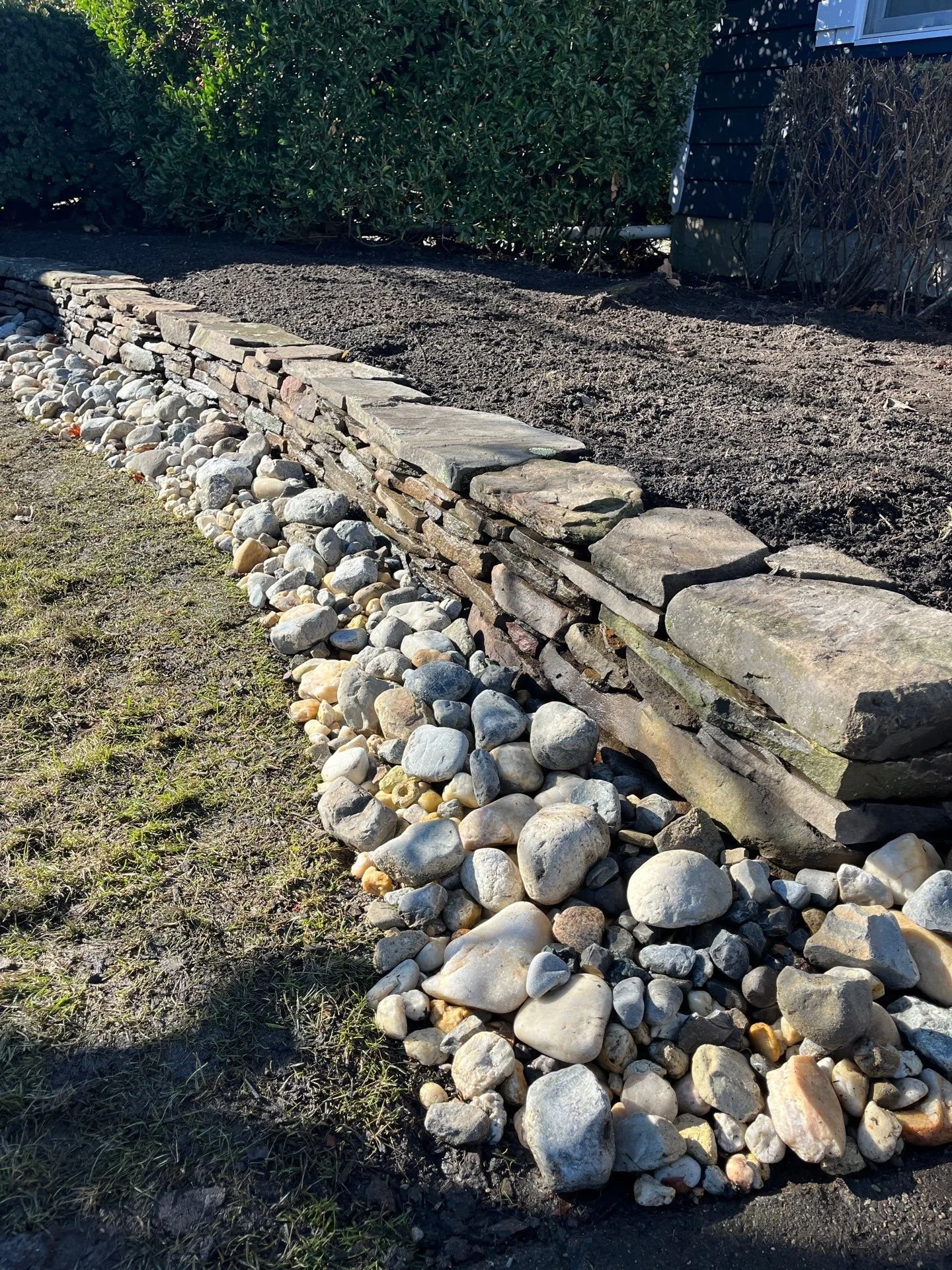 Low dry-stacked natural stone wall with a river rock border edging a garden bed at a Rumson, NJ home.