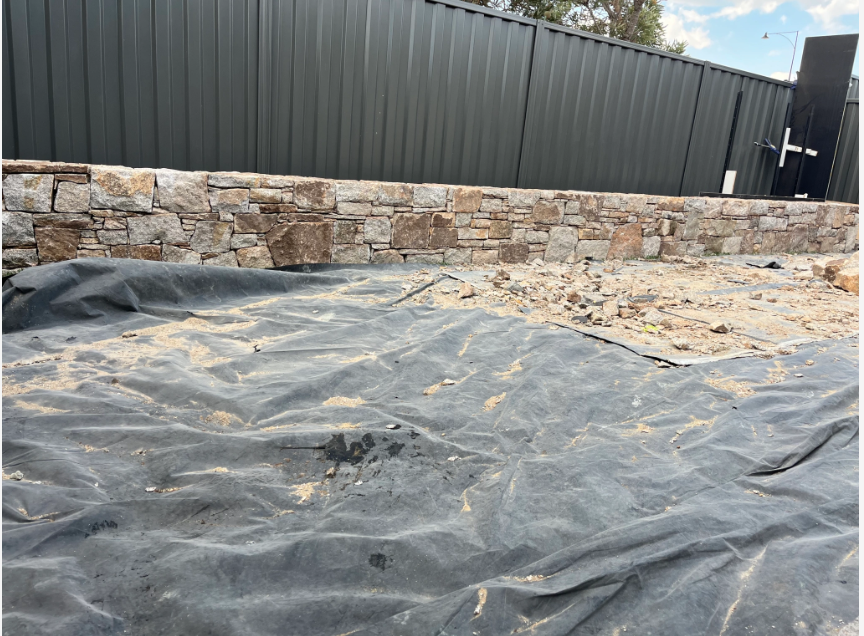 View of a construction site with black tarp on the ground and a stone wall in front of a metal fence.