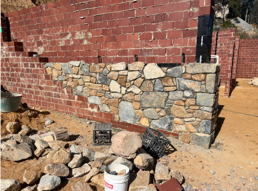 Construction site with a half-built stone and brick wall next to a sandy ground. Several stones and building materials are scattered around, along with a plastic bucket and black crates.