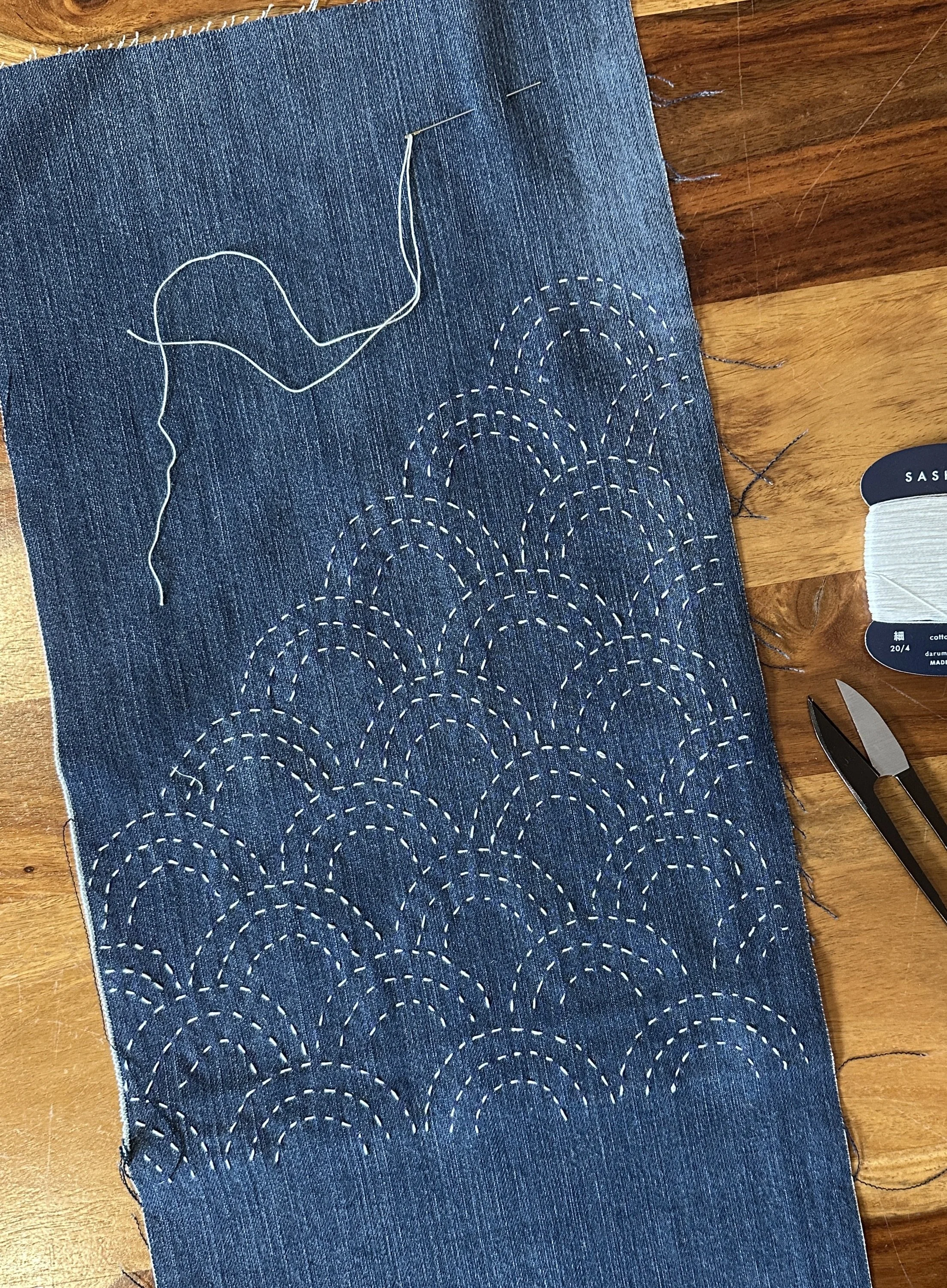 A piece of dark blue denim fabric with white dashed embroidery stitching in a wave pattern. Sewing pins are visible at the top edge, and a pair of scissors is on the right edge of the fabric, which is placed on a wooden surface.