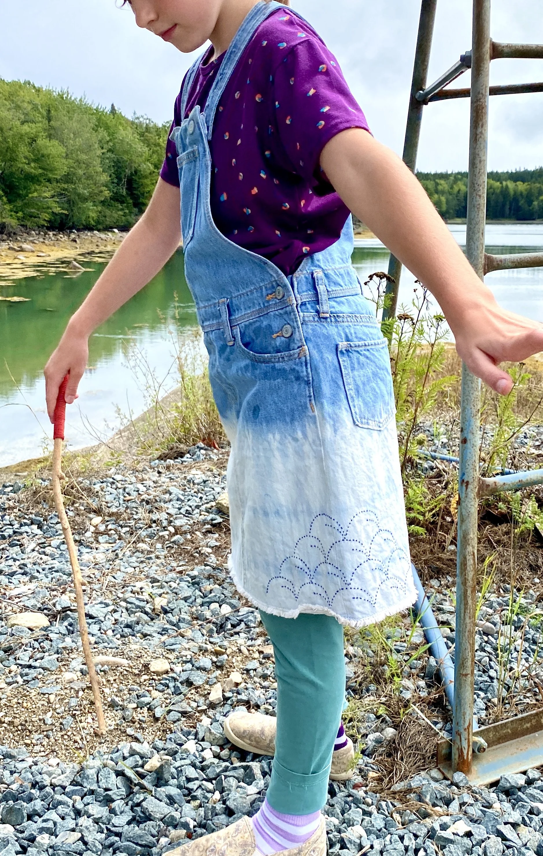 A child wearing teal pants, striped socks, a purple shirt with sprinkles, denim overalls, holding a walking stick, standing outdoors near a rocky shoreline with water and trees in the background.