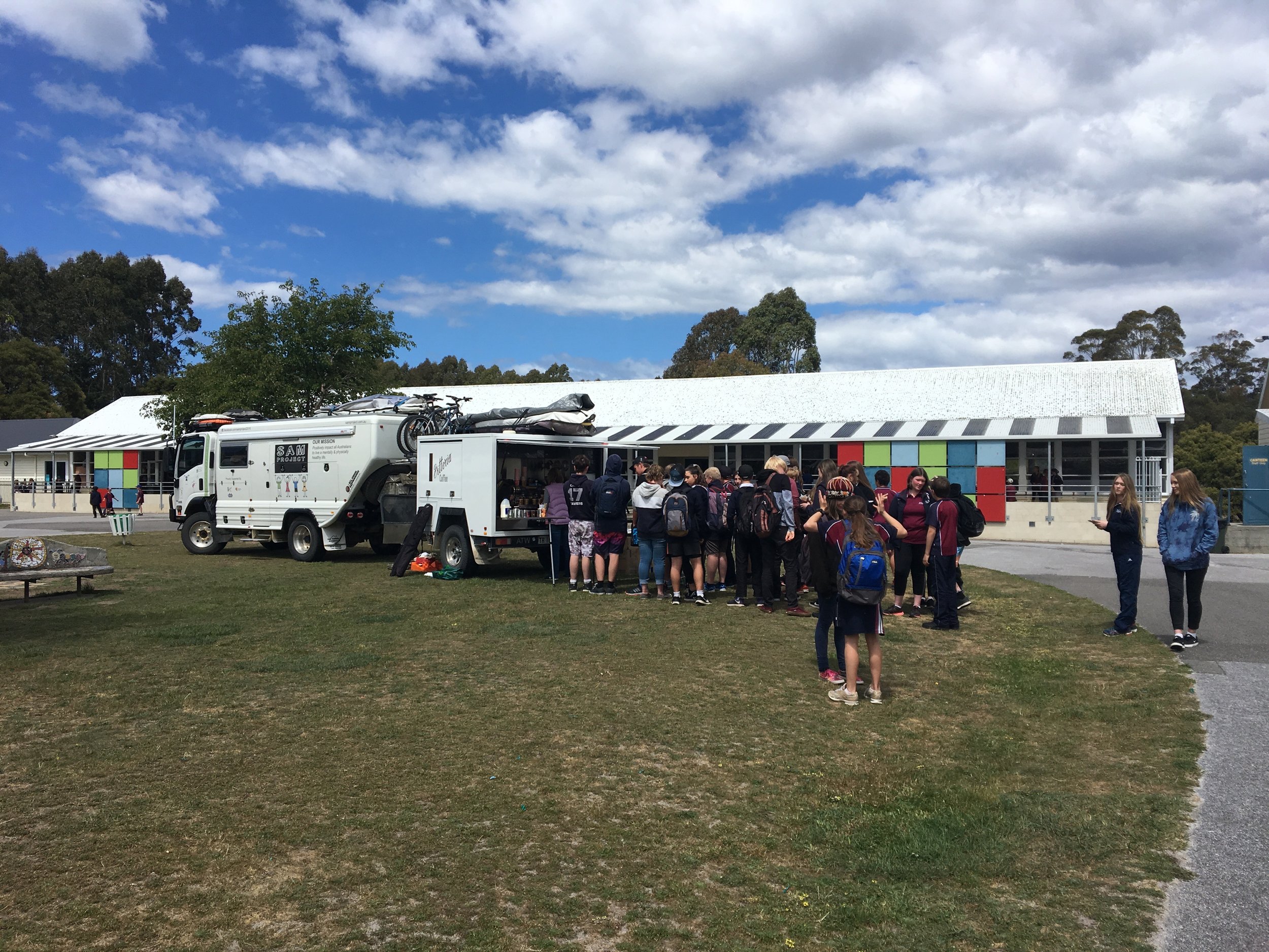 A group of students waiting in line at a food truck in front of a school building on a partly cloudy day.