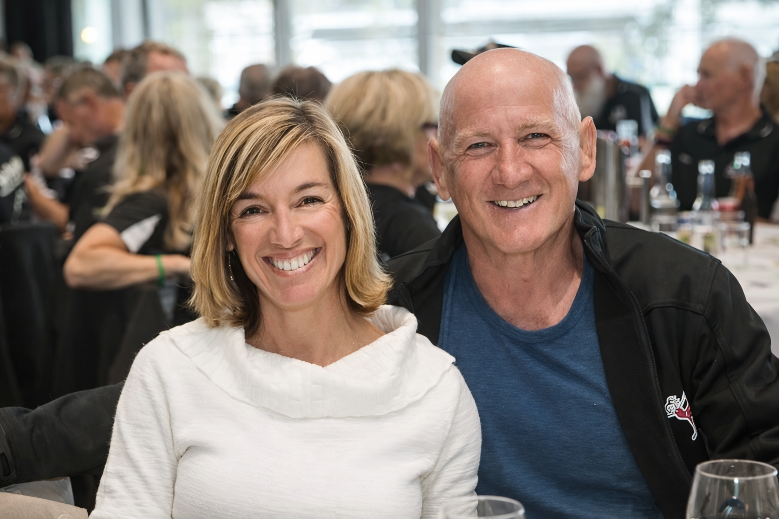 A smiling woman with shoulder-length blonde hair and a man with a shaved head, both sitting together at a social event or gathering. The woman wears a white top, and the man wears a blue t-shirt and a black jacket. There are other people in the background at a table with drinks, enjoying the event.