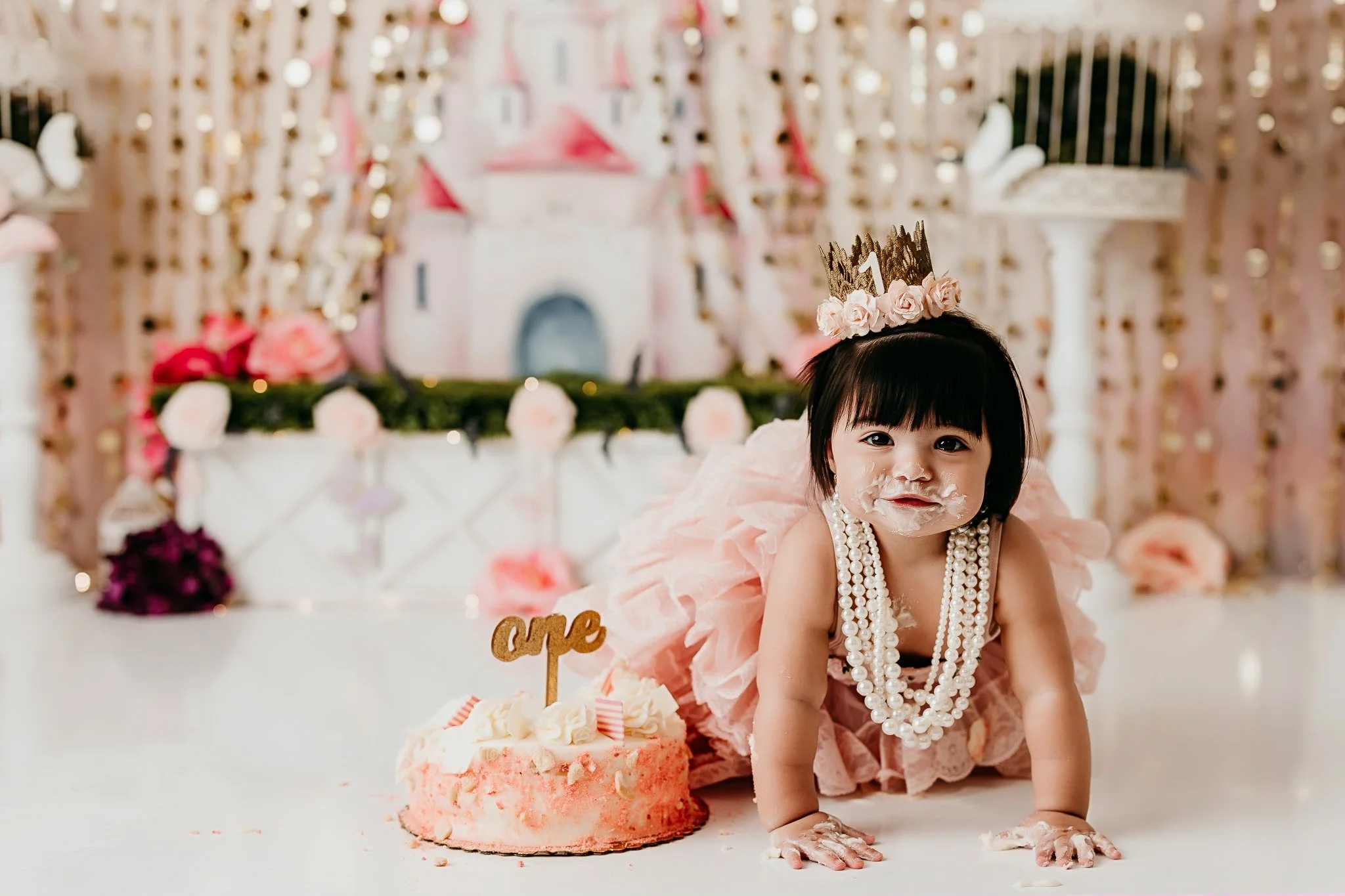 A baby wearing pink in a tiara eating a cake.