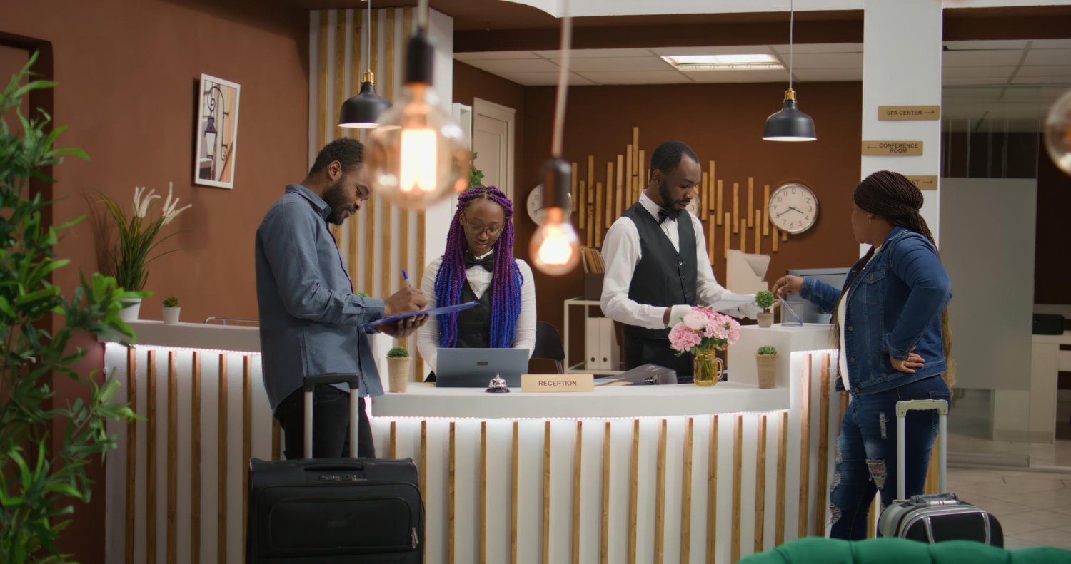 A hotel reception area with three staff members behind the counter and a guest with a suitcase in front of it. The staff includes one man and one woman, both in black vests and shirts, and another woman with purple braids wearing a white shirt. The guest is a woman with braids wearing a denim jacket and jeans, holding a card or key. The reception desk has potted plants and a sign that reads 'RECEPTION.' The background shows a wall with clocks and directional signs to spa and conference rooms, decorated with wooden accents and hanging lamps.